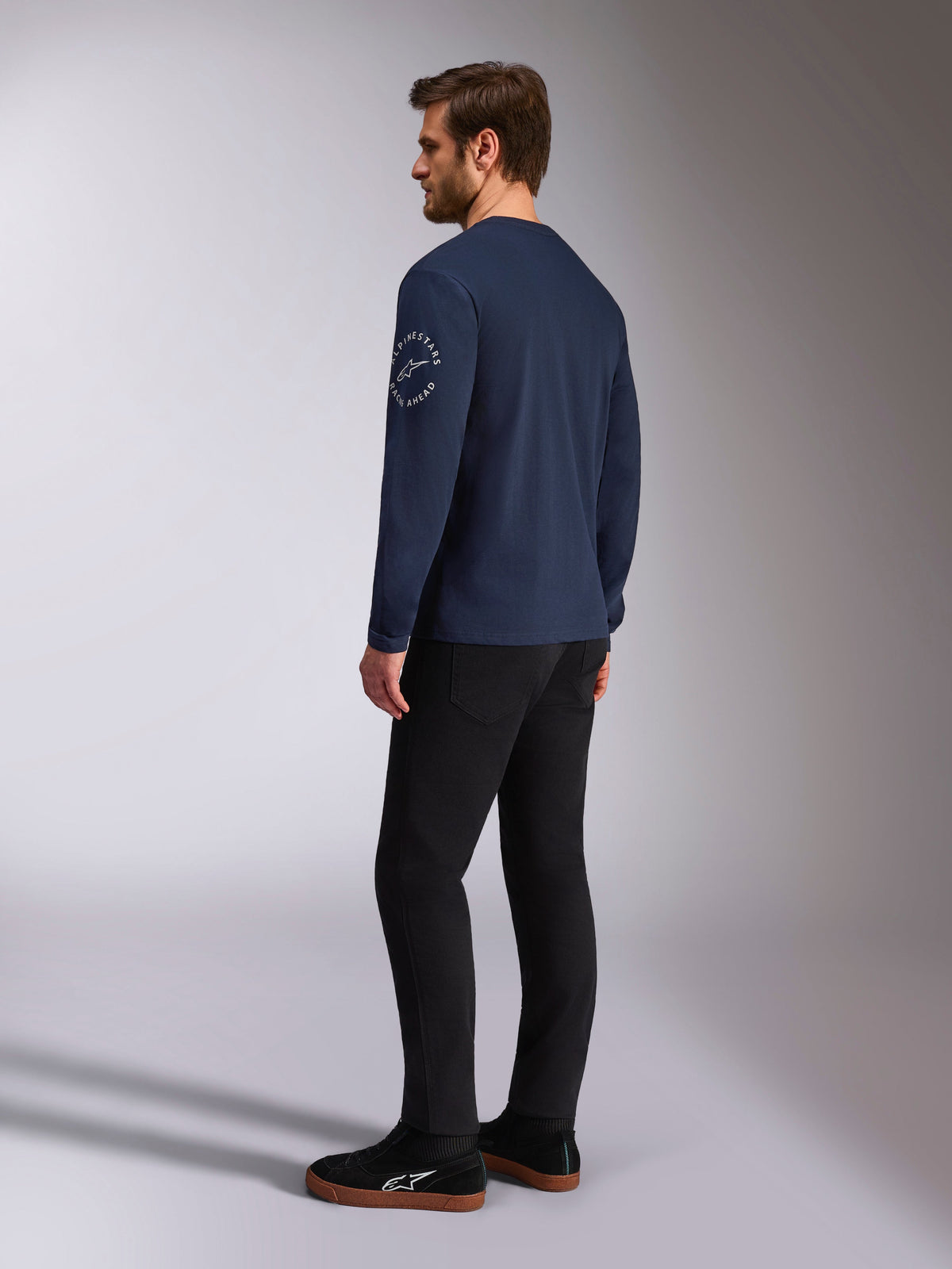 Alpinestars Mandatory CSF langærmet t-shirt, navyblå, hvidt cirkulært logo på ærmet, set bagfra fra siden, kombineret med sorte bukser og sorte sneakers, casual motorsport-streetwear