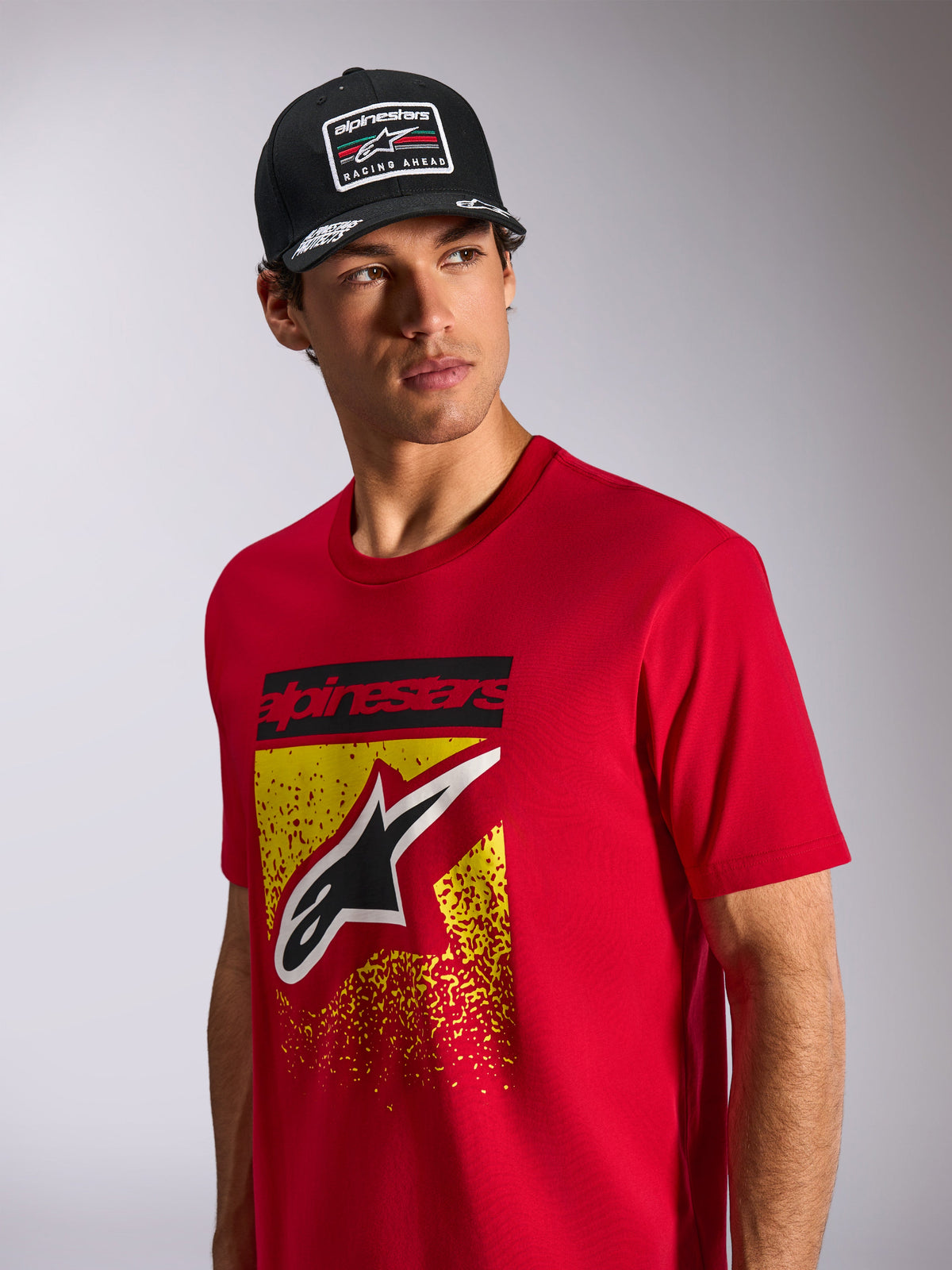 Alpinestars Noka CSF Tee, kortærmet t-shirt, rød, med Astars-logo på fronten med gul og rød MX-graduering, båret med en sort Alpinestars 'Racing Ahead' kasket, casual motorsport-streetwear