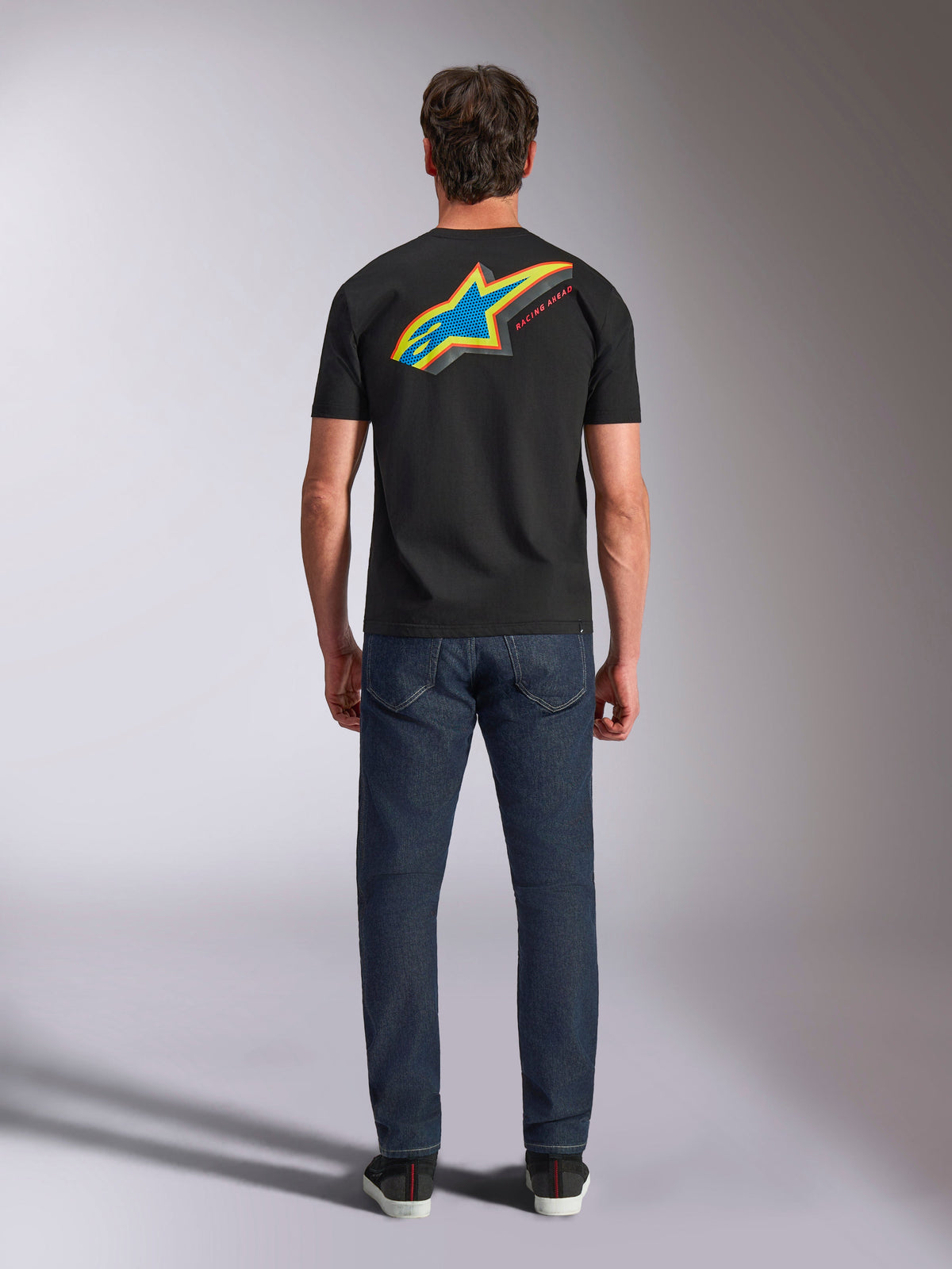 Alpinestars Angler CSF Tee, kortærmet T-shirt, sort, model set bagfra iført trøje med stort livligt flerfarvet Astars-stjernelogo og 'RACING AHEAD'-tekst, kombineret med mørkeblå jeans, afslappet motorsport-streetwear