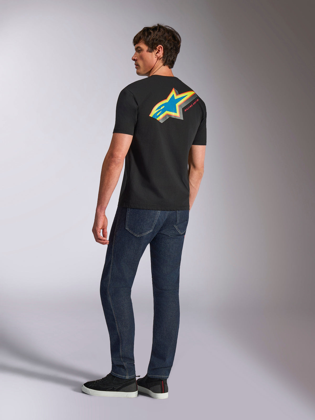Alpinestars Angler CSF Tee, kortærmet t-shirt, sort, med et stort, farverigt Astars-logo på ryggen med blå, gule og røde perforerede detaljer, båret med mørkeblå jeans, afslappet motorsport streetwear