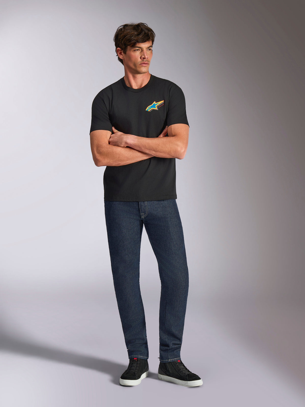 Alpinestars Angler CSF Tee, kortærmet T-shirt, sort, livligt flerfarvet Astars-logo på brystet, båret af model med korslagte arme, mørke denimjeans og sorte sneakers, afslappet motorsport streetwear