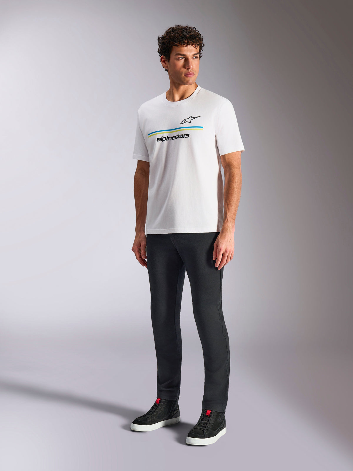 Alpinestars Lanes CSF Tee, hvid kortærmet T-shirt, båret af model med sorte slim-fit bukser og sorte high-top sneakers, med blå og gule vandrette striber og Alpinestars-branding på brystet, afslappet motorsport-inspireret streetwear