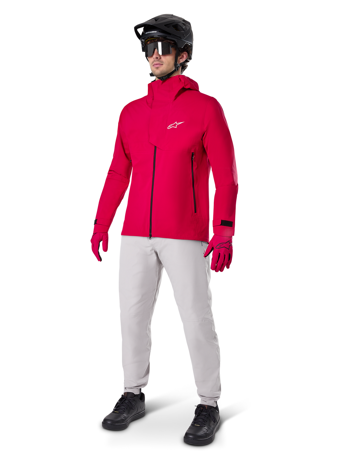 Mandlig cyklist iført Alpinestars A-Dura Elite vandtæt jakke i Red Fluo, kombineret med lysegrå tekniske bukser, røde handsker, sort MTB-hjelm med goggles og sorte sko, komplet højtydende outfit til mountainbiking