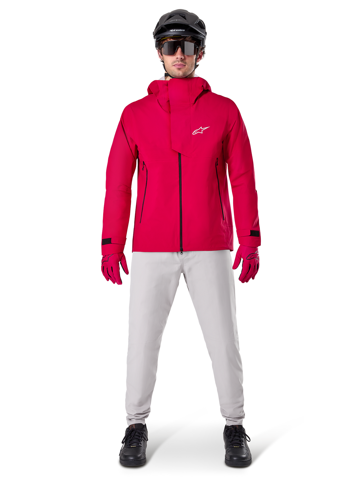 Alpinestars A-Dura Elite vandtæt jakke, Red Fluo, teknisk mountainbike-jakke båret med en sort MTB-hjelm, solbriller, matchende røde handsker og lysegrå bukser, fuldkropsvisning af cykeludstyr til alle vejrforhold