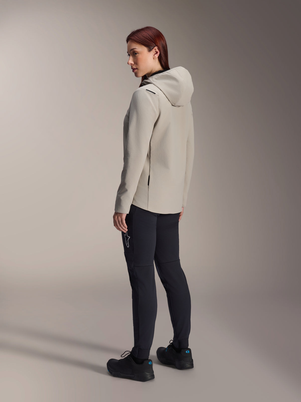 Alpinestars Stella A-Dura Thermal Jacket i Titanium Gray, trekvart visning bagfra af en kvinde iført den isolerede cykeljakke med hætte og sorte MTB-bukser, studiebillede der viser den regulære pasform og lommedetaljer.