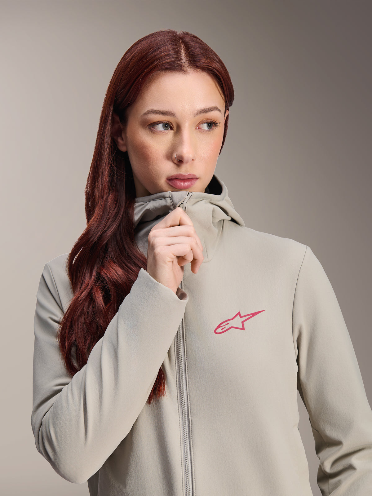 Alpinestars Stella A-Dura termojakke til kvinder, titaniumgrå med lyserødt Alpinestars-stjernelogo, model med langt rødt hår, der bærer og lyner den hætteklædte jakke, letvægtsisoleret cykeltøj til koldt vejr