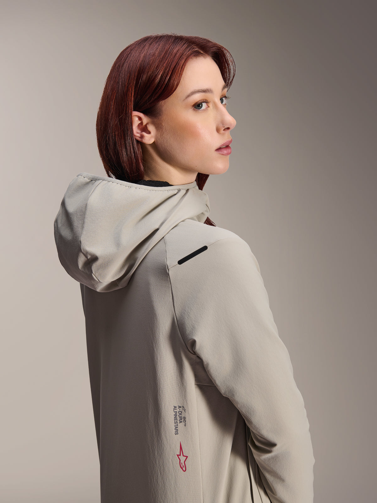 Alpinestars Stella A-Dura Thermal Jacket, MTB-jakke til kvinder, Titanium grå, kvinde der kigger over sin skulder iført performance-jakke med hætte, med Alpinestars-tekst og rødt stjernelogo på siden, designet til cykling i koldt vejr