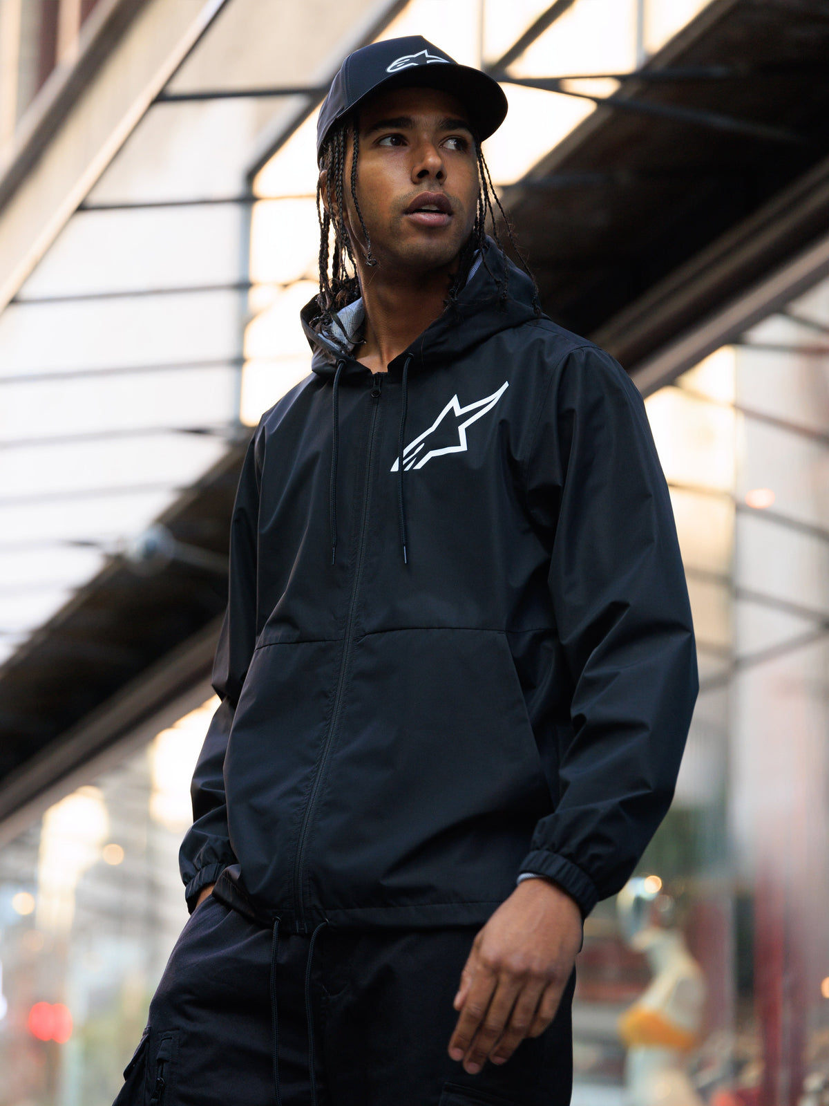 Alpinestars Speeder Chromium Windbreak Jakke, sort vindjakke med hætte og hvidt logo på brystet, båret af en mand med en matchende sort Alpinestars kasket i et urbant miljø, afslappet motorsport streetwear.