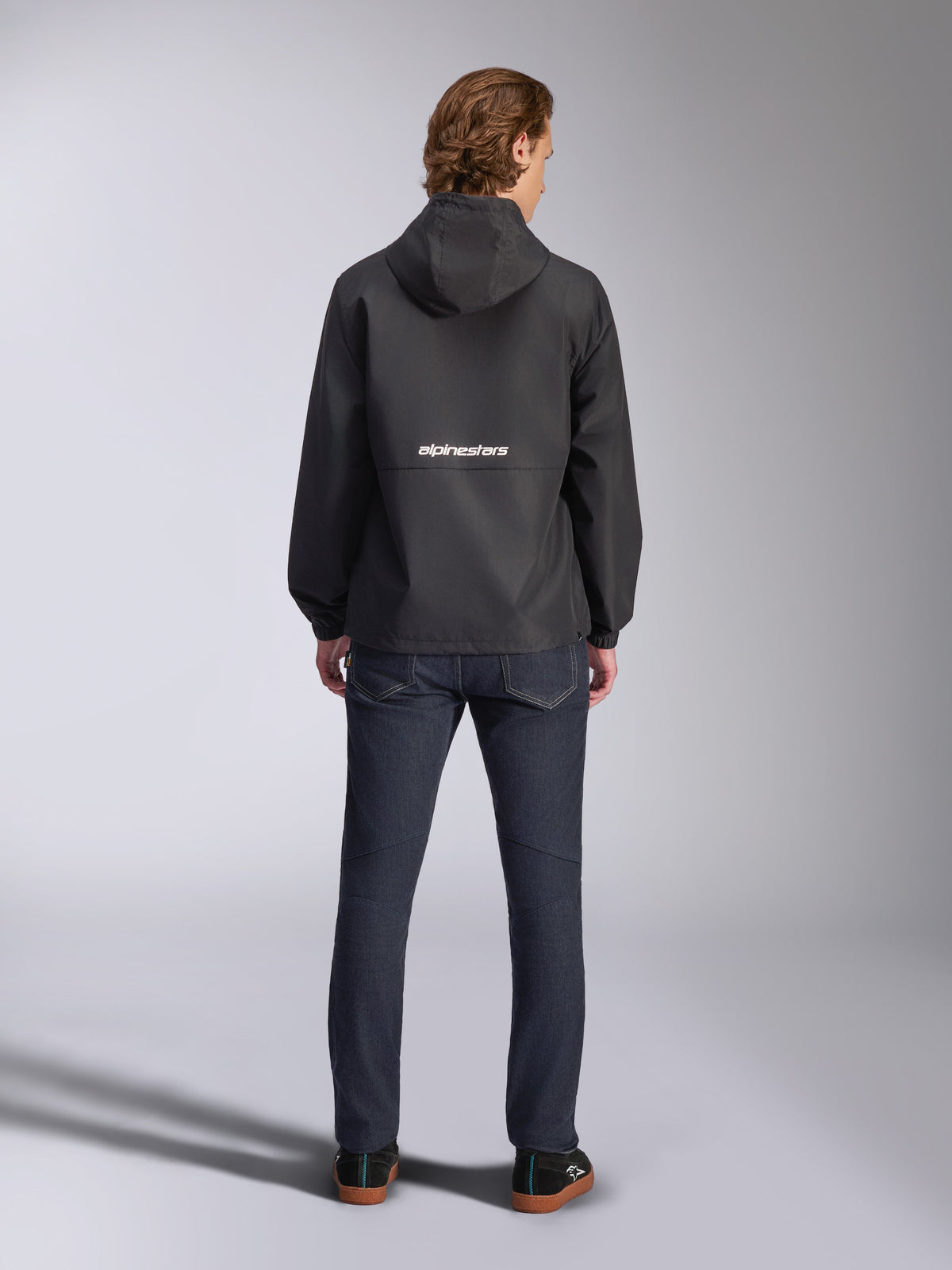 Alpinestars Speeder Chromium Windbreaker-jakke, sort, set bagfra med hvid Alpinestars-branding på tværs af midten, design med hætte, båret af model med mørke denimbukser, afslappet motorsport-inspireret jakke