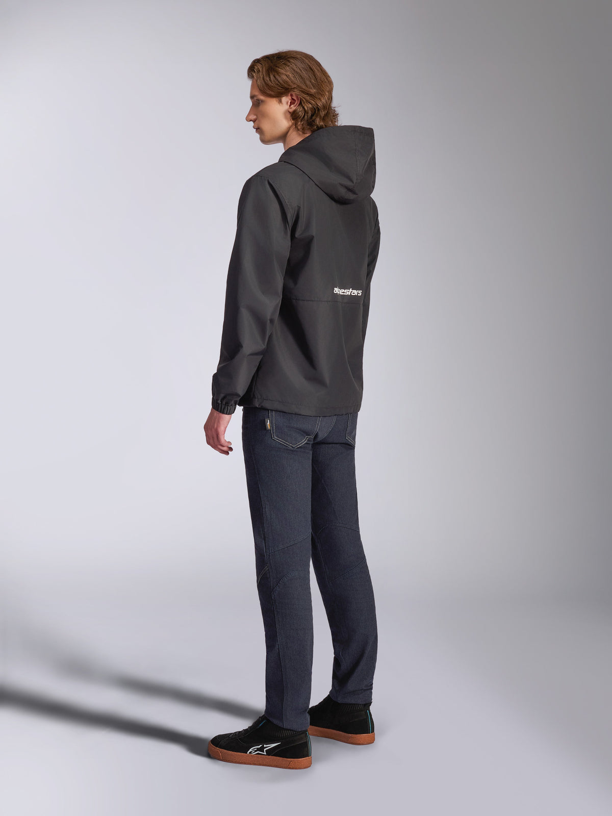 Alpinestars Speeder Chromium Windbreaker-jakke, sort med hvidt logo på ryggen, casual vindjakke til mænd med hætte, set fra siden båret med mørke jeans og sorte sneakers, motorsportsinspireret streetwear