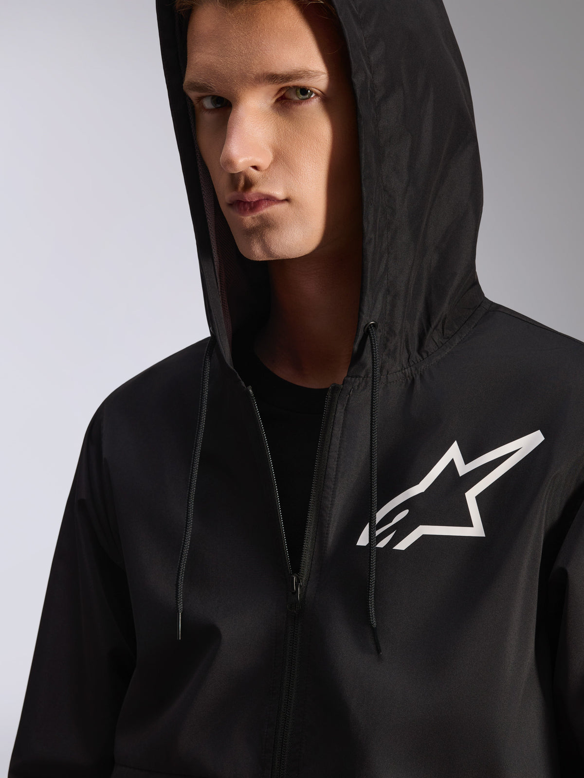 Alpinestars Speeder Chromium Windbreaker Jakke, sort, nærbillede af en windbreaker med hætte og et hvidt stjernelogo på brystet, har en hætte med snoretræk, letvægts tekstilmateriale, afslappet motorsport-inspireret streetwear