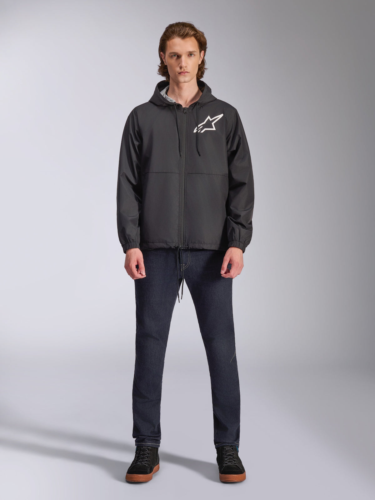 Alpinestars Speeder Chromium Windbreaker-jakke, sort med hvidt Astars-logo, bæres af model med mørkeblå jeans og sorte sneakers, design med hætte og lynlås foran, afslappet motorsport-inspireret streetwear