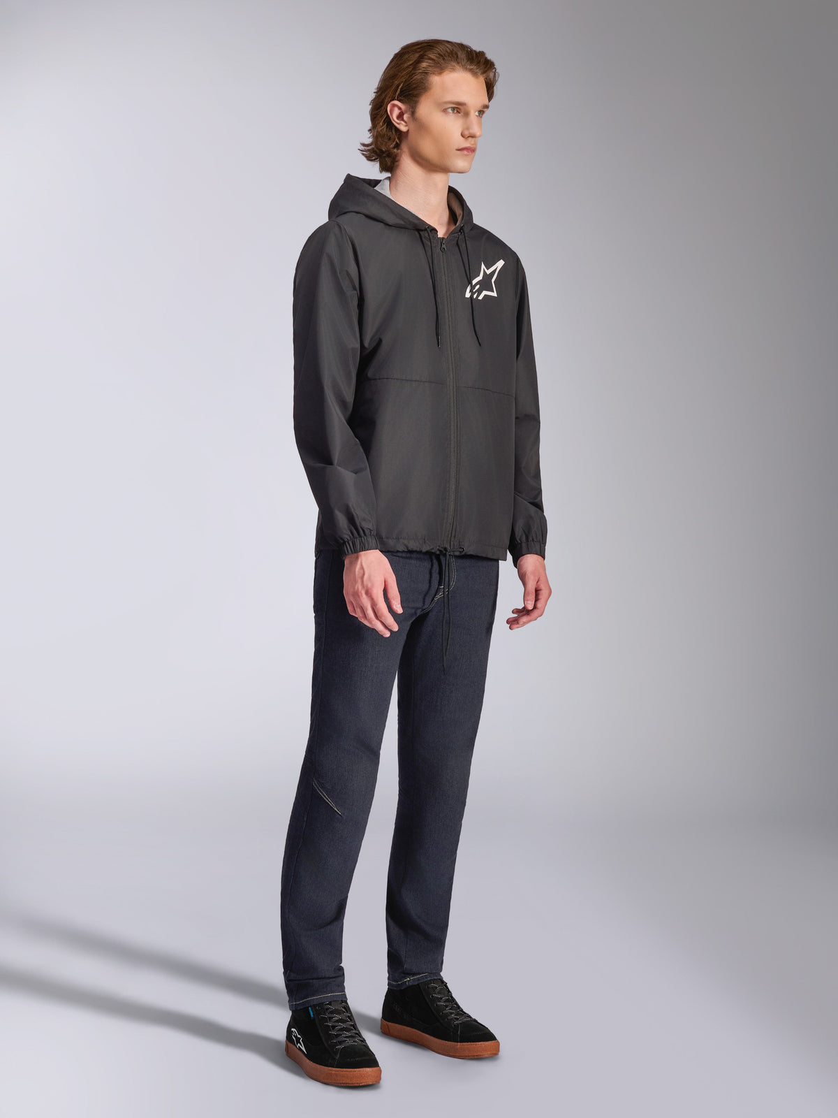 Alpinestars Speeder Chromium Windbreaker Jakke, sort, windbreaker med hætte og lynlås foran, hvidt Alpinestars stjerne-logo på venstre bryst, afslappet motorsport-streetwear, stylet med mørke bukser og sorte sneakers i et studie