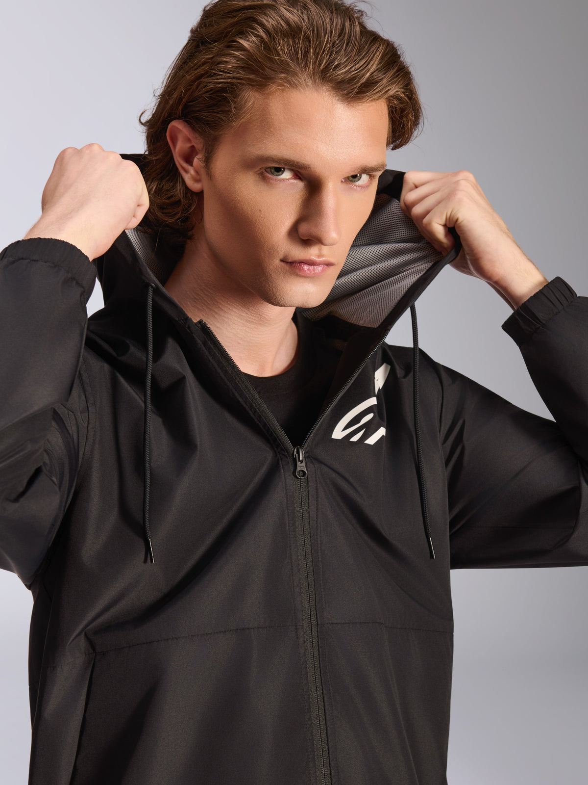 Alpinestars Speeder Chromium Windbreaker-jakke, sort, bæres af en mandlig model, der holder i hætten, har et hvidt logo på brystet, mesh-foring i hætten og gennemgående lynlås foran, afslappet motorsport-inspireret streetwear