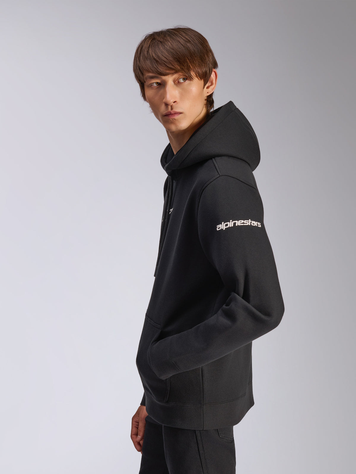 Alpinestars Frontal hættetrøje, sort pullover-fleece, model set fra siden, hvidt Alpinestars-navnetræk på ærmet, kængurulomme, afslappet motorsport-inspireret streetwear