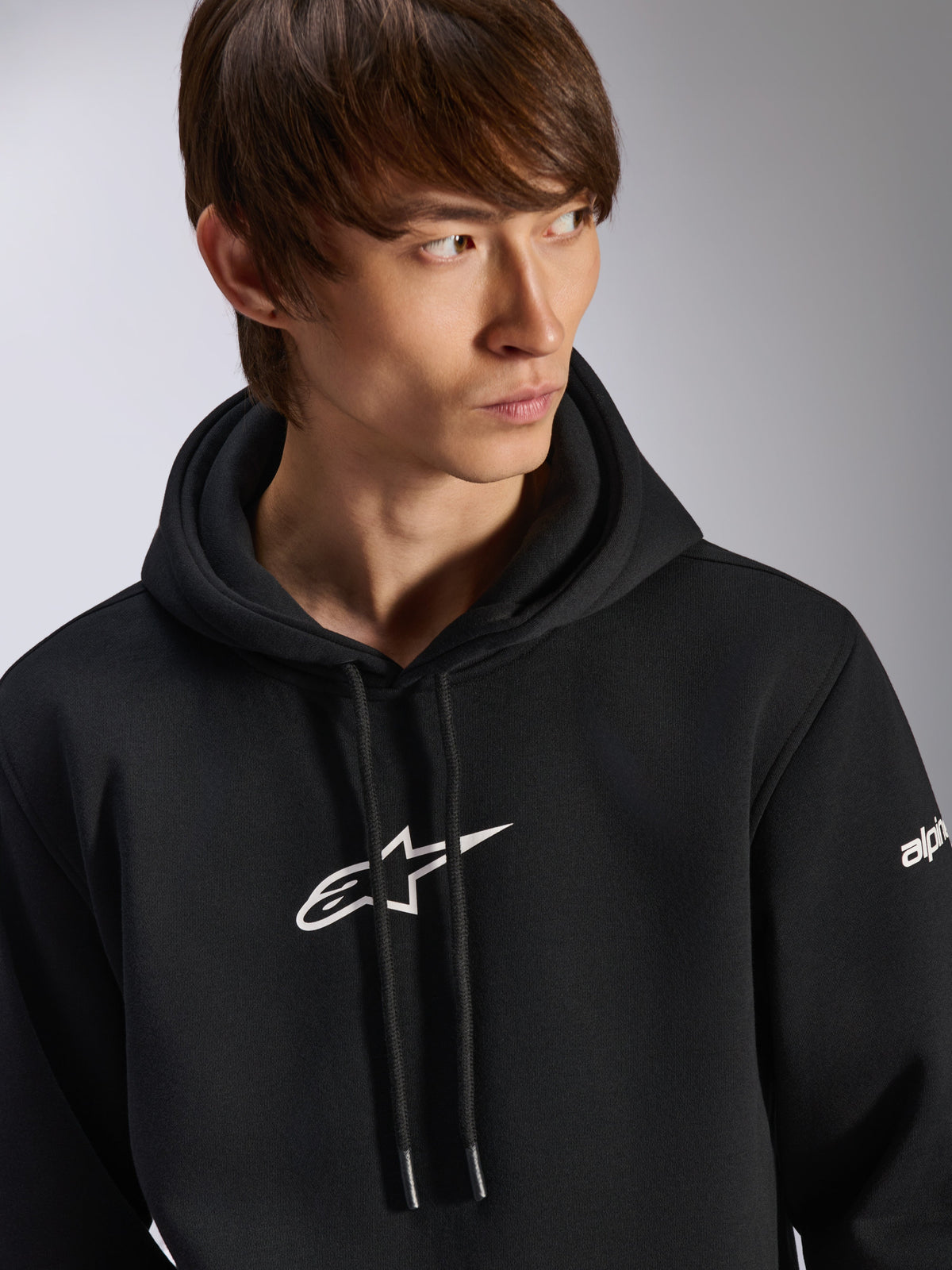 Alpinestars Frontal hættetrøje, sort pullover fleece-hættetrøje, hvidt Astar-logo på brystet og wordmark-branding på ærmet, hætte med snoretræk, båret af mandlig model, afslappet motorsport-streetwear