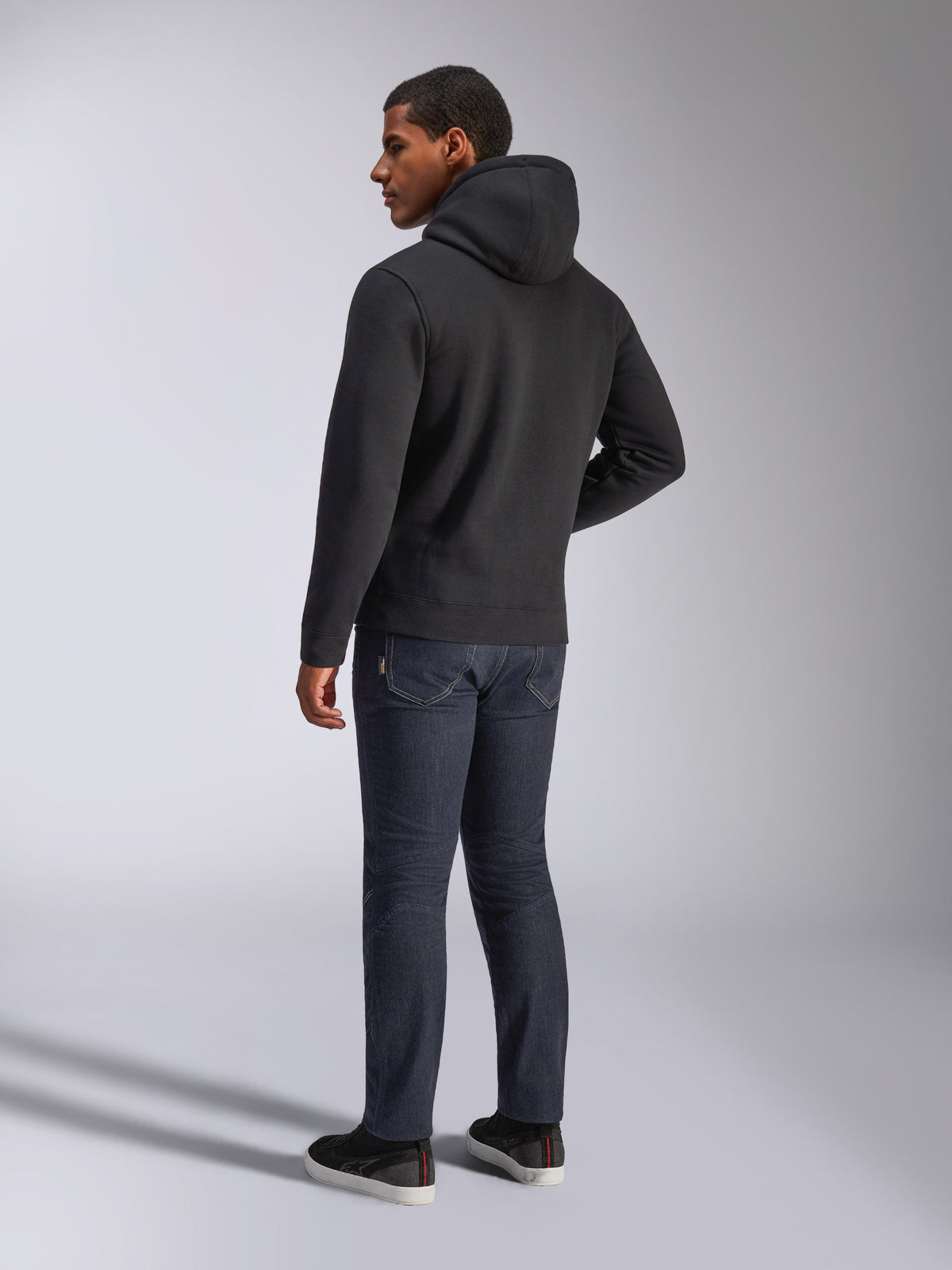 Alpinestars Blaze V3 hættetrøje, set bagfra på en mand iført den sorte pullover-hættetrøje, kombineret med mørke jeans og sorte sneakers, afslappet motorsport-inspireret streetwear
