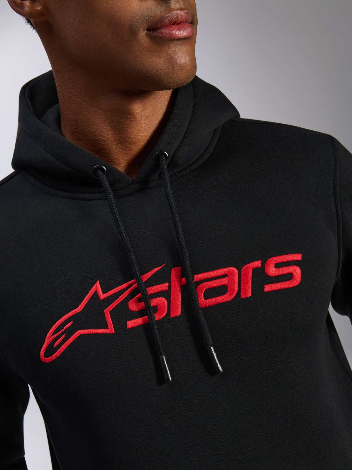 Alpinestars Blaze V3 hættetrøje, nærbillede af brystet, sort fleecestof med rødt lineært Alpinestars-logoprint, hætte med snøre og dyppede ender, afslappet motorsport-inspireret streetwear