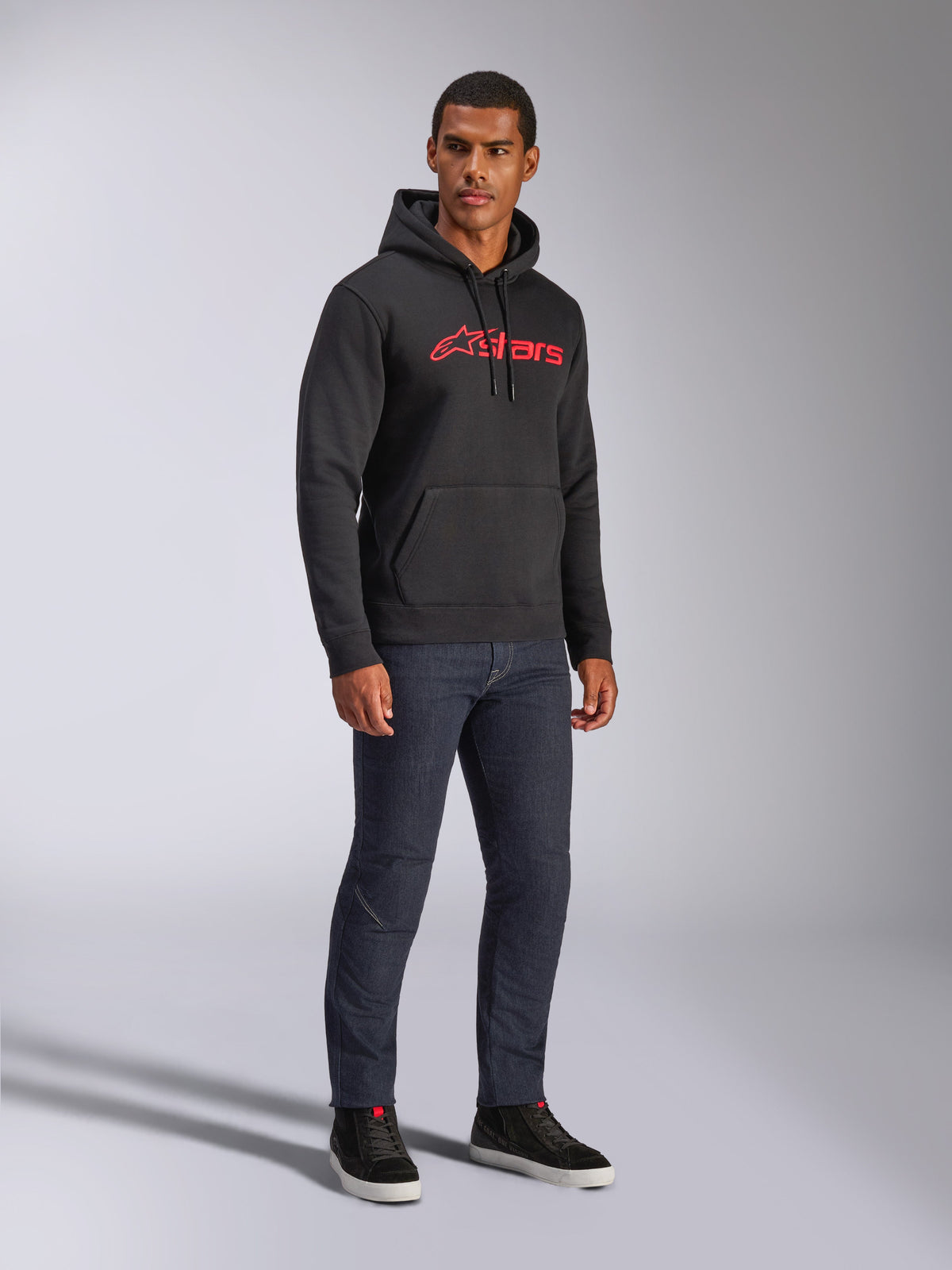 Alpinestars Blaze V3 hættetrøje, sort pullover-fleece med rødt Alpinestars-logo på brystet, båret af mandlig model med mørke denim-jeans og sorte sneakers, afslappet motorsport-inspireret streetwear