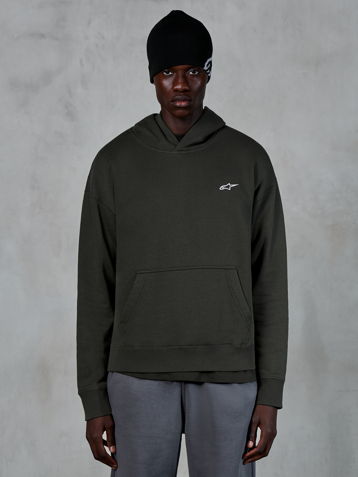 Alpinestars Essentials Ageless Hoodie, casual streetwear, Off Black pullover med et lille hvidt Alpinestars-logo på brystet, båret med en sort beanie, minimalistisk design med kængurulomme og stenvasket finish