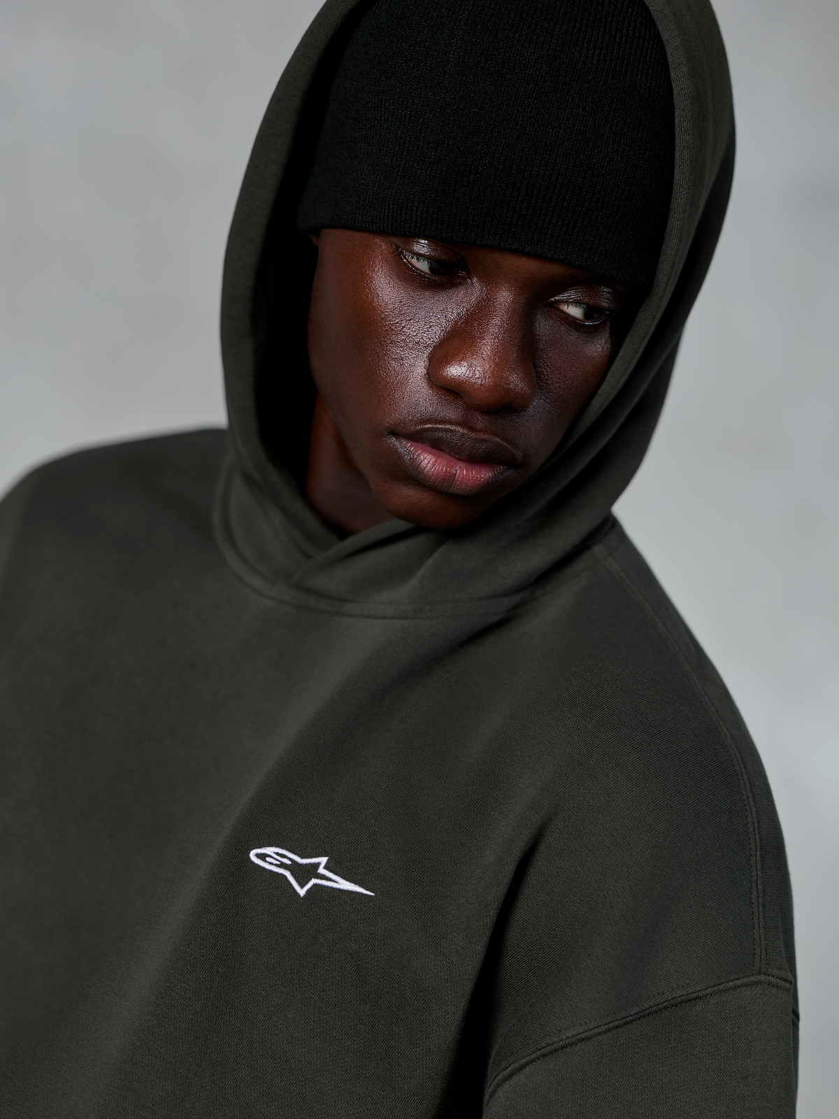 Alpinestars Essentials Ageless Hoodie, Off Black, nærbillede af model med en sort hue under hætten, lille hvidt stjernelogo på brystet, afslappet motorsport-inspireret streetwear