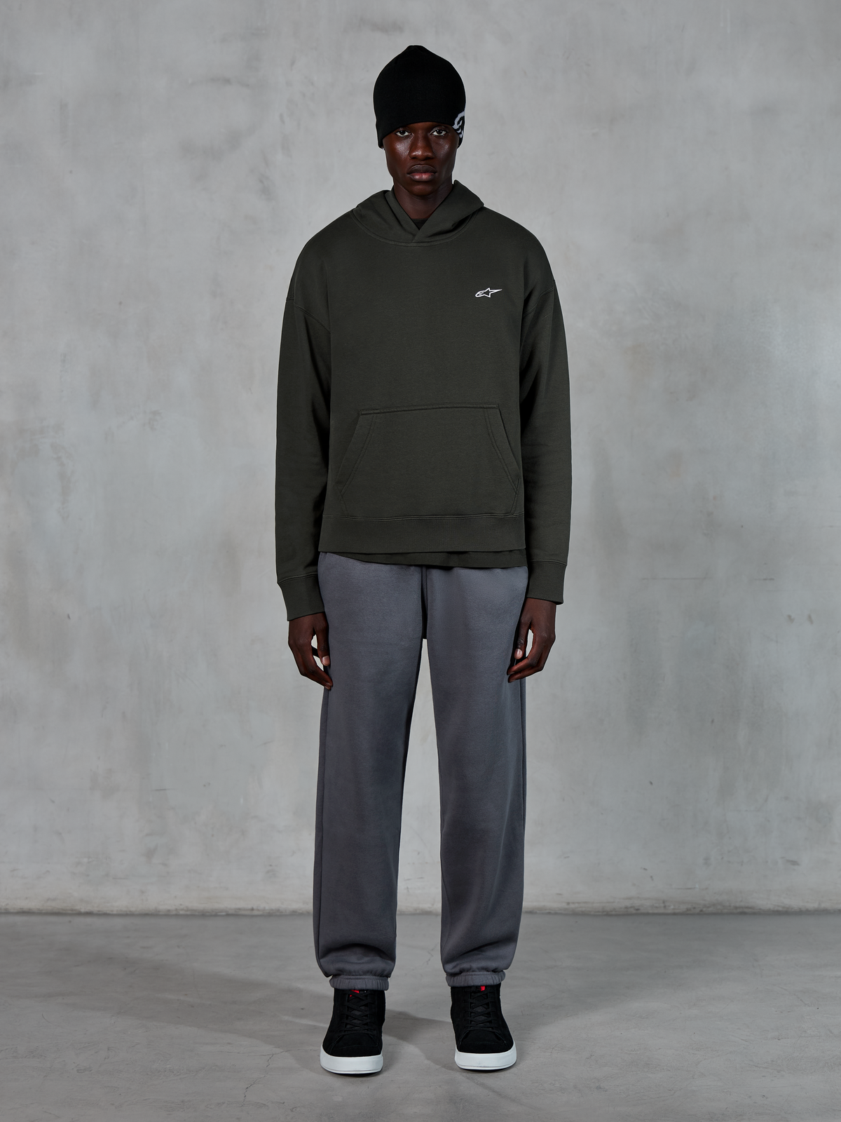 Model iført Alpinestars Essentials Ageless Hoodie, afslappet streetwear, Off Black med lille hvidt logo på brystet, kombineret med grå joggingbukser, sort beanie og sorte sneakers, minimalistisk motorsport-inspireret design