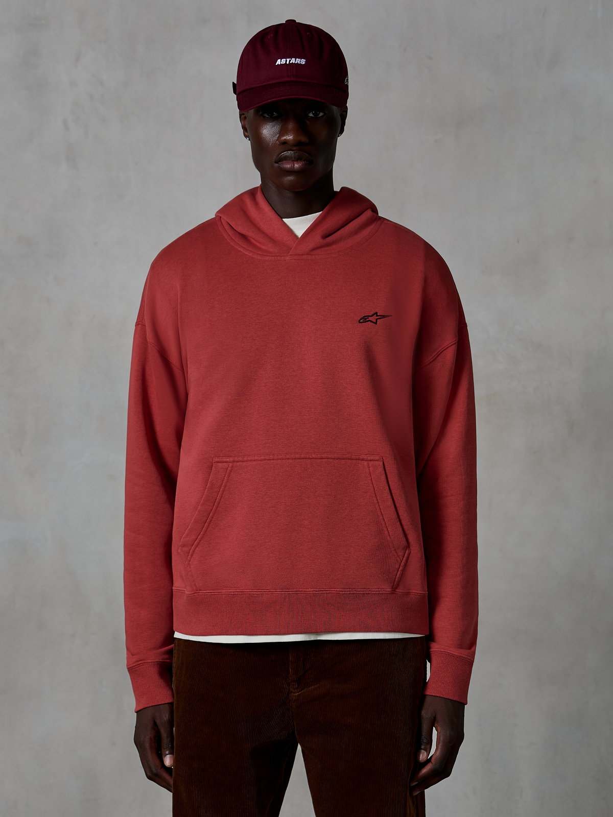 Alpinestars Essentials Ageless hættetrøje, casual streetwear, teglrød farve, lille sort stjernelogo på brystet, kængurulomme, stenvasket finish, bæres af en model med bordeaux kasket og brune fløjlsbukser