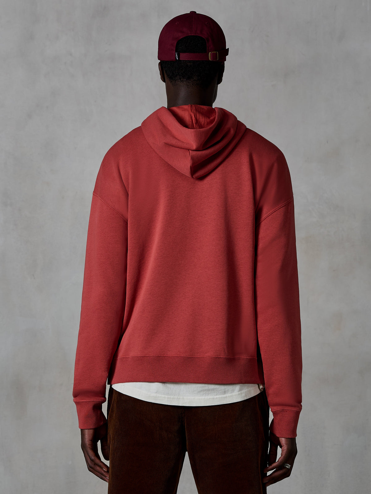 Alpinestars Essentials Ageless Hoodie, afslappet fleece-hættetrøje, teglrød, set bagfra af model iført livsstilstøj, enkelt design med ribkant forneden og ved manchetterne, stylet med brune fløjlsbukser og en bordeaux kasket