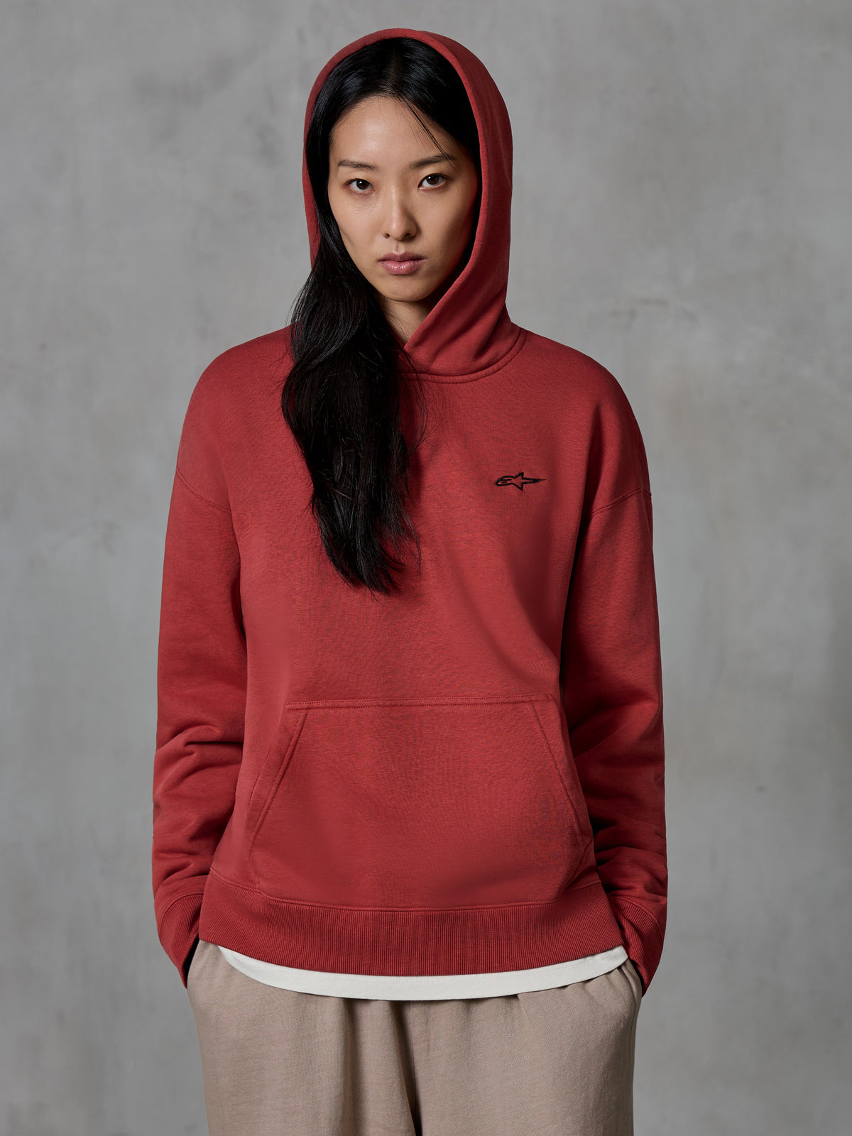 Alpinestars Essentials Ageless Hoodie, teglrød pullover sweatshirt, båret af en kvindelig model med hætten oppe, lille sort broderet stjernelogo på brystet, kængurulomme, afslappet motorsport-inspireret lifestyle streetwear