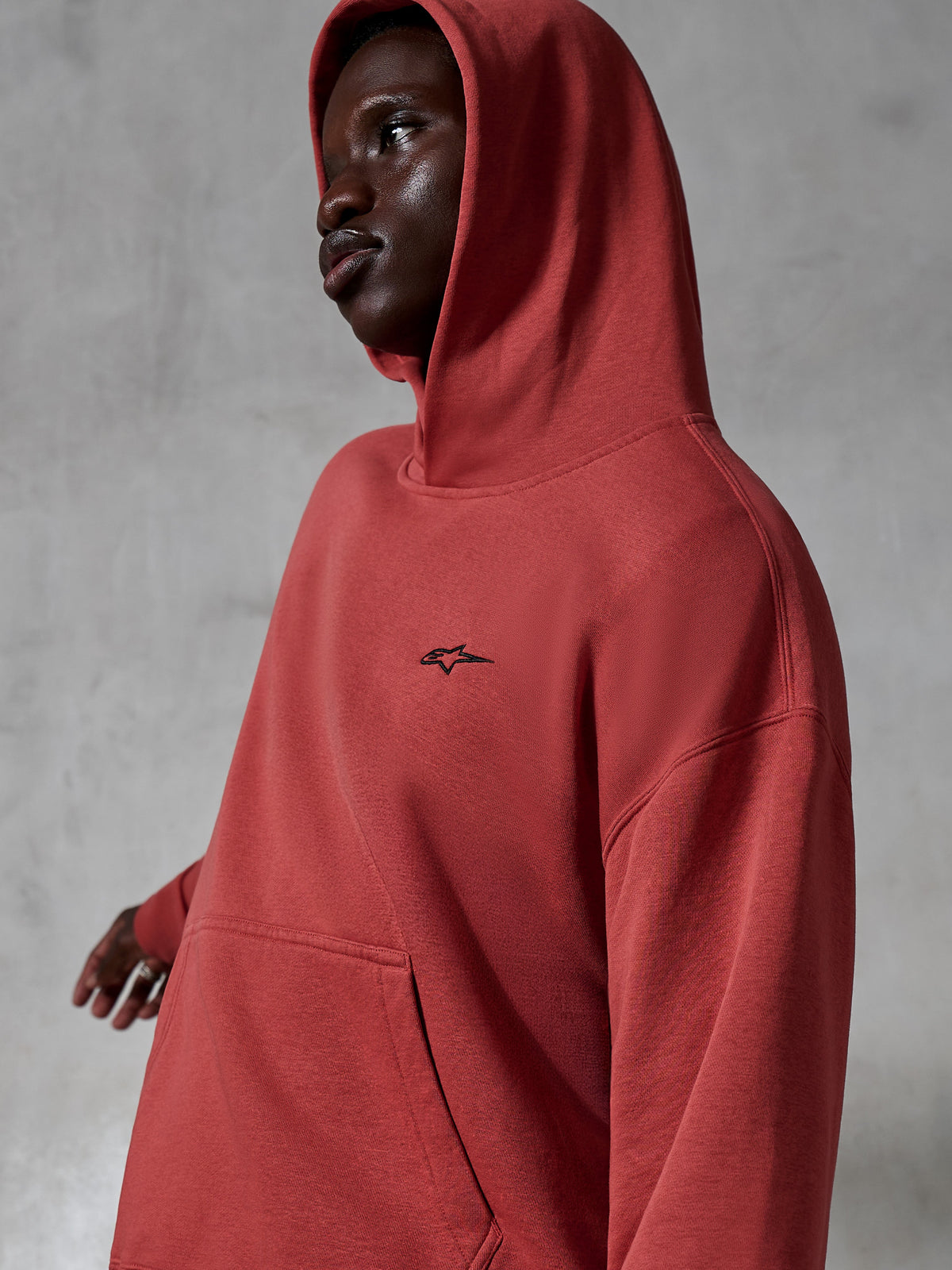 Alpinestars Essentials Ageless hættetrøje, afslappet sportstøj, teglrød, bæres af en model med hætten oppe, lille sort broderet logo, blød fleece-tekstur, afslappet pasform streetwear