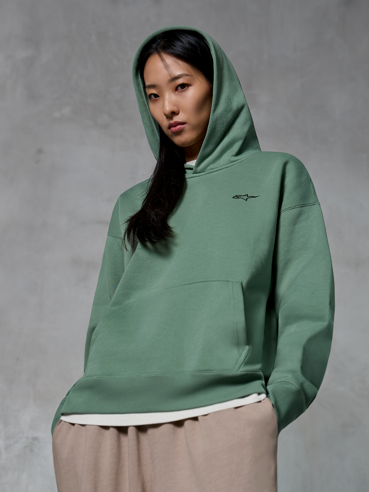 Alpinestars Essentials Ageless Hoodie, afslappet streetwear, Cactus-grøn med lille sort logo, hættetrøje båret med hætten oppe, kængurulomme foran, stenvasket finish, kombineret med lysebrune bukser