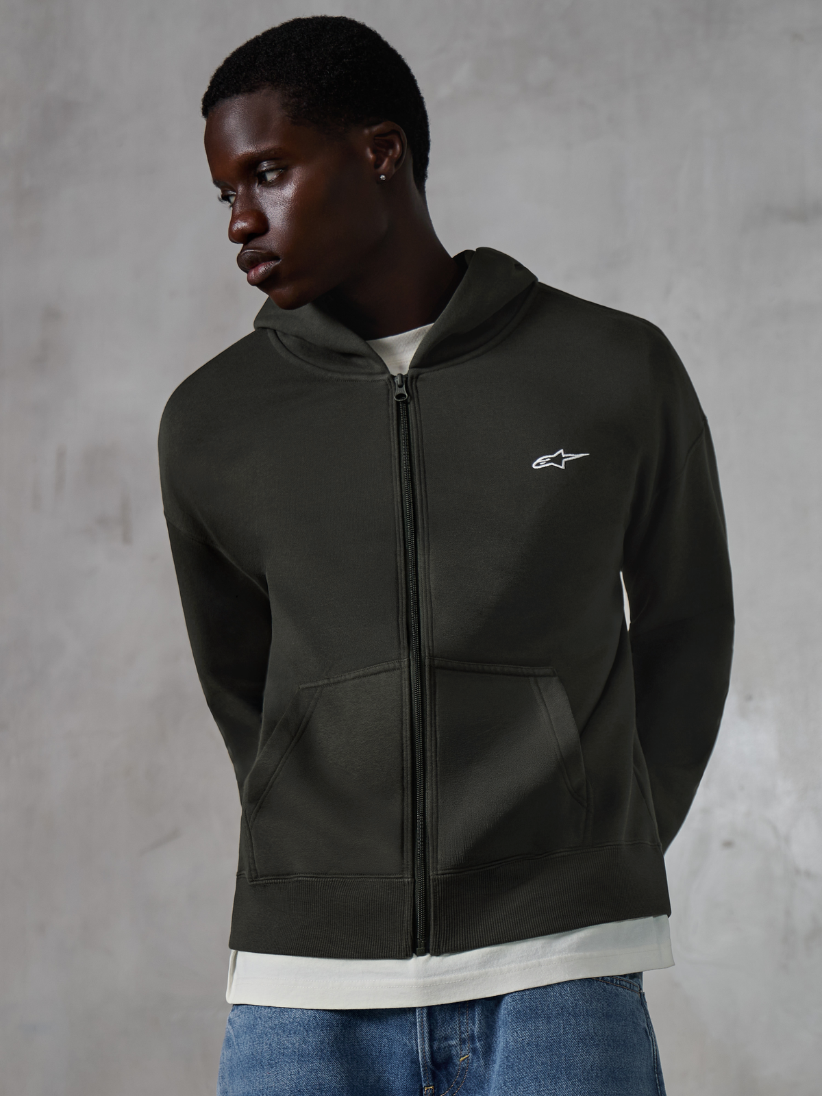 Alpinestars Essentials Ageless Zip Hoodie, Off Black casual streetwear, fleece med gennemgående lynlås, lille hvidt Alpinestars-logo på brystet, båret af en model med blå jeans, afslappet pasform med forlommer og stone-washed finish.