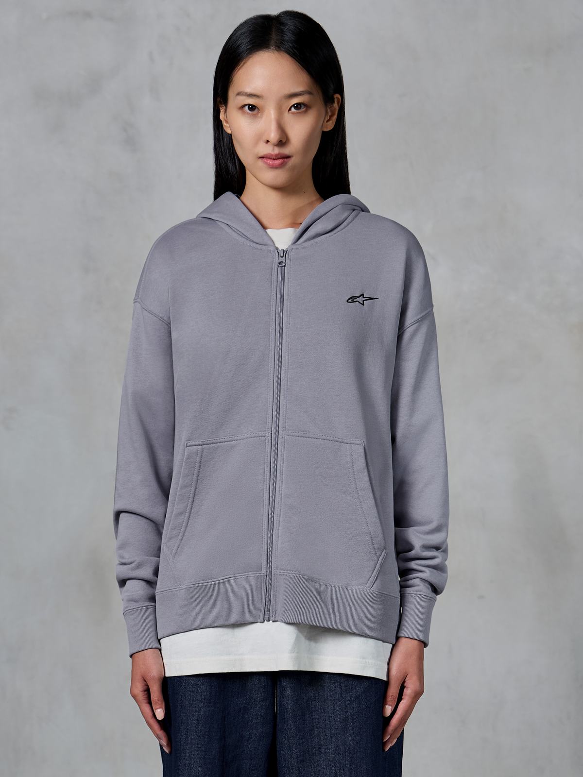 Alpinestars Essentials Ageless Zip Hoodie, Silver, afslappet streetwear, fleece-sweatshirt med gennemgående lynlås, lille sort Alpinestars-logo på brystet, delt kængurulomme, bæres af model over hvid t-shirt og mørke jeans