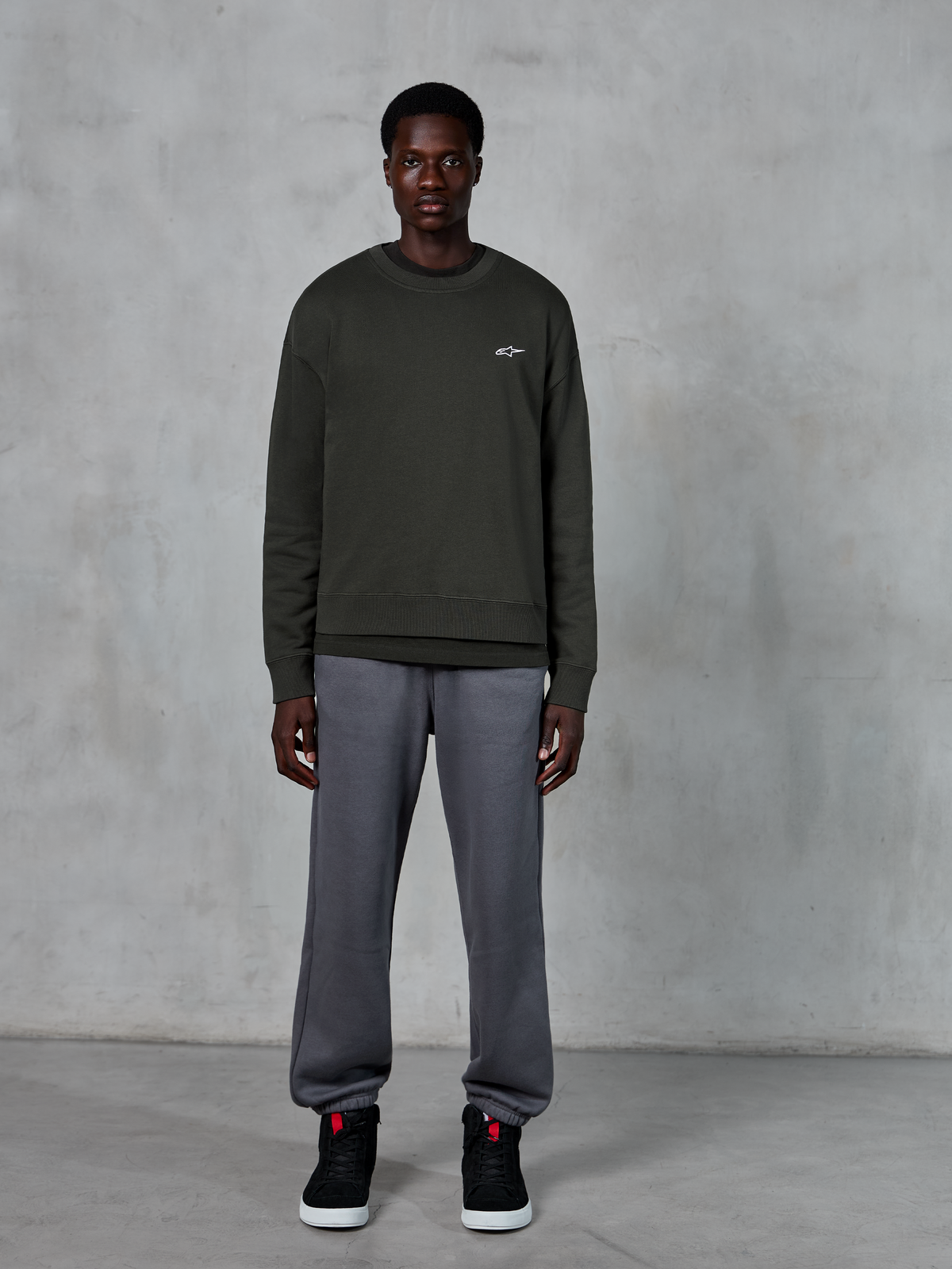 Alpinestars Essentials Ageless Crew, mørkegrøn crewneck sweatshirt med et lille hvidt logo på brystet, båret af en model med grå joggingbukser og sorte high-top sneakers, afslappet livsstilstøj.
