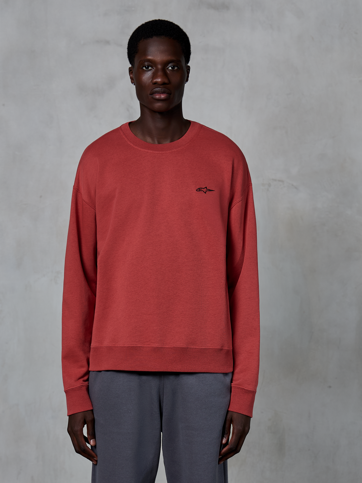 Alpinestars Essentials Ageless Crew, crewneck sweatshirt, teglrød, lille sort logo på brystet, båret af en model med grå bukser, premium blød fleece, afslappet streetwear
