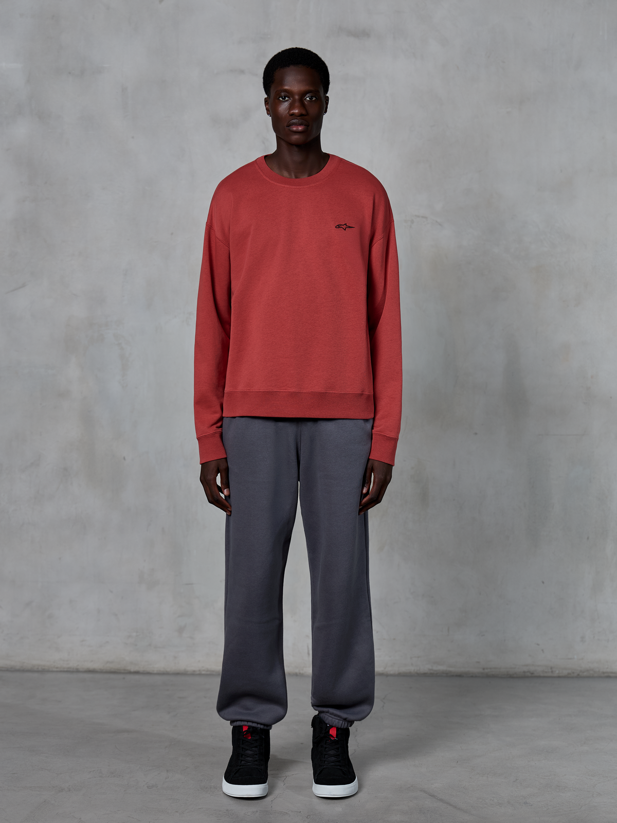 Alpinestars Essentials Ageless Crew, teglrød crewneck sweatshirt, båret med grå joggingbukser og sorte sneakers, med et lille sort logo på brystet, afslappet motorsport-inspireret streetwear