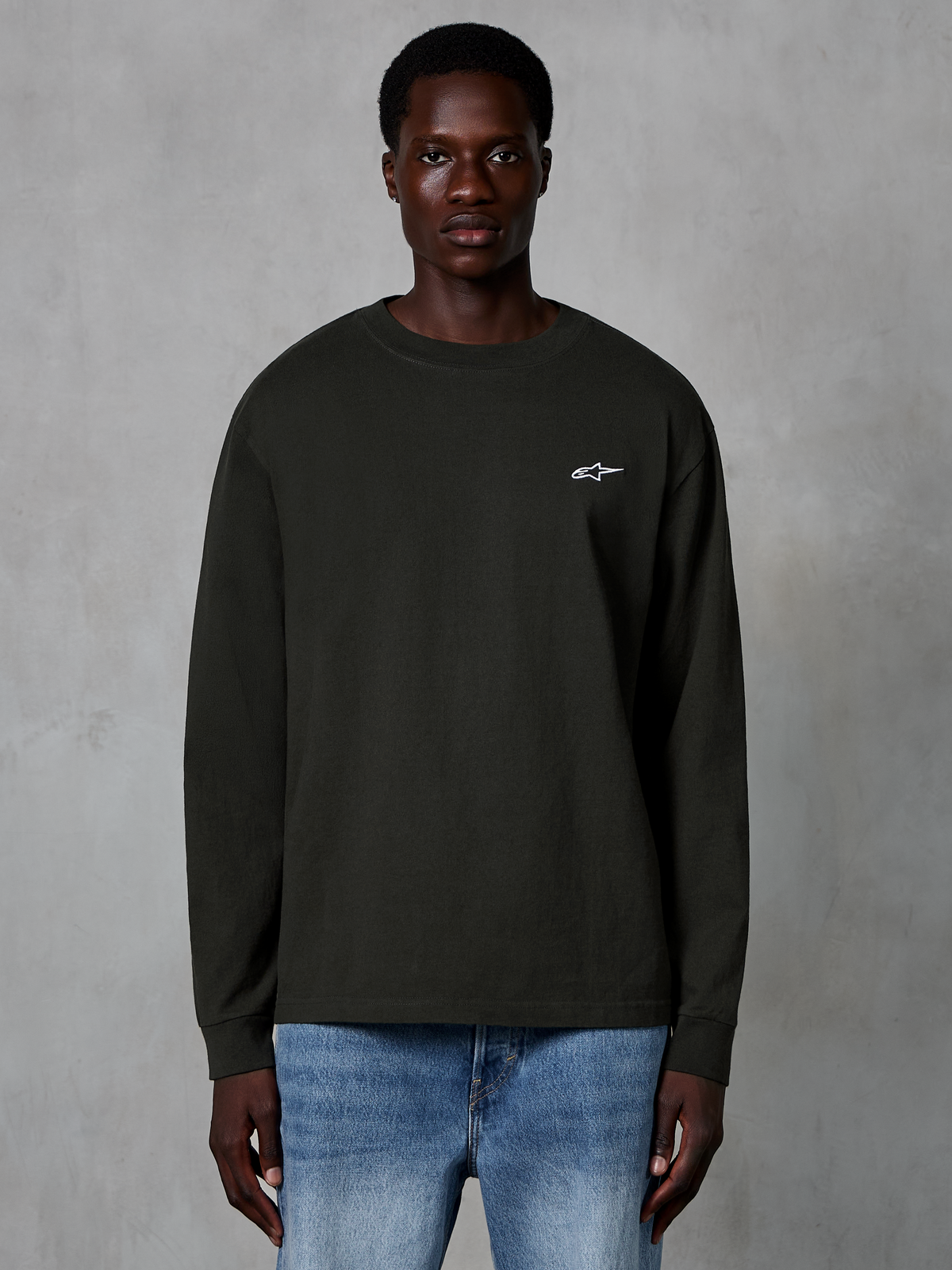 Alpinestars Essentials Ageless LS Tee, langærmet t-shirt, Off Black, med et lille hvidt Alpinestars-logo på brystet, båret af en model med blå jeans, afslappet streetwear i økologisk bomuld