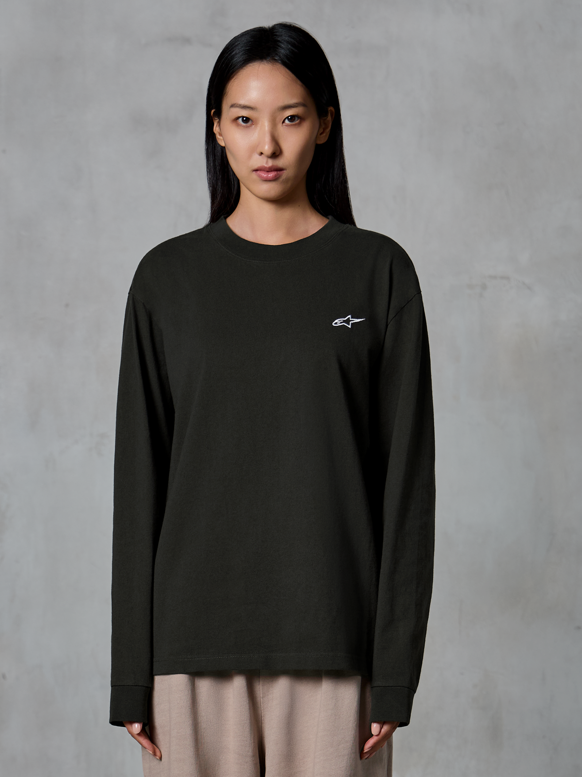 Model iført Alpinestars Essentials Ageless LS Tee, langærmet T-shirt, Off Black, med et lille hvidt Alpinestars-logo på brystet, kombineret med beige bukser, afslappet lifestyle streetwear