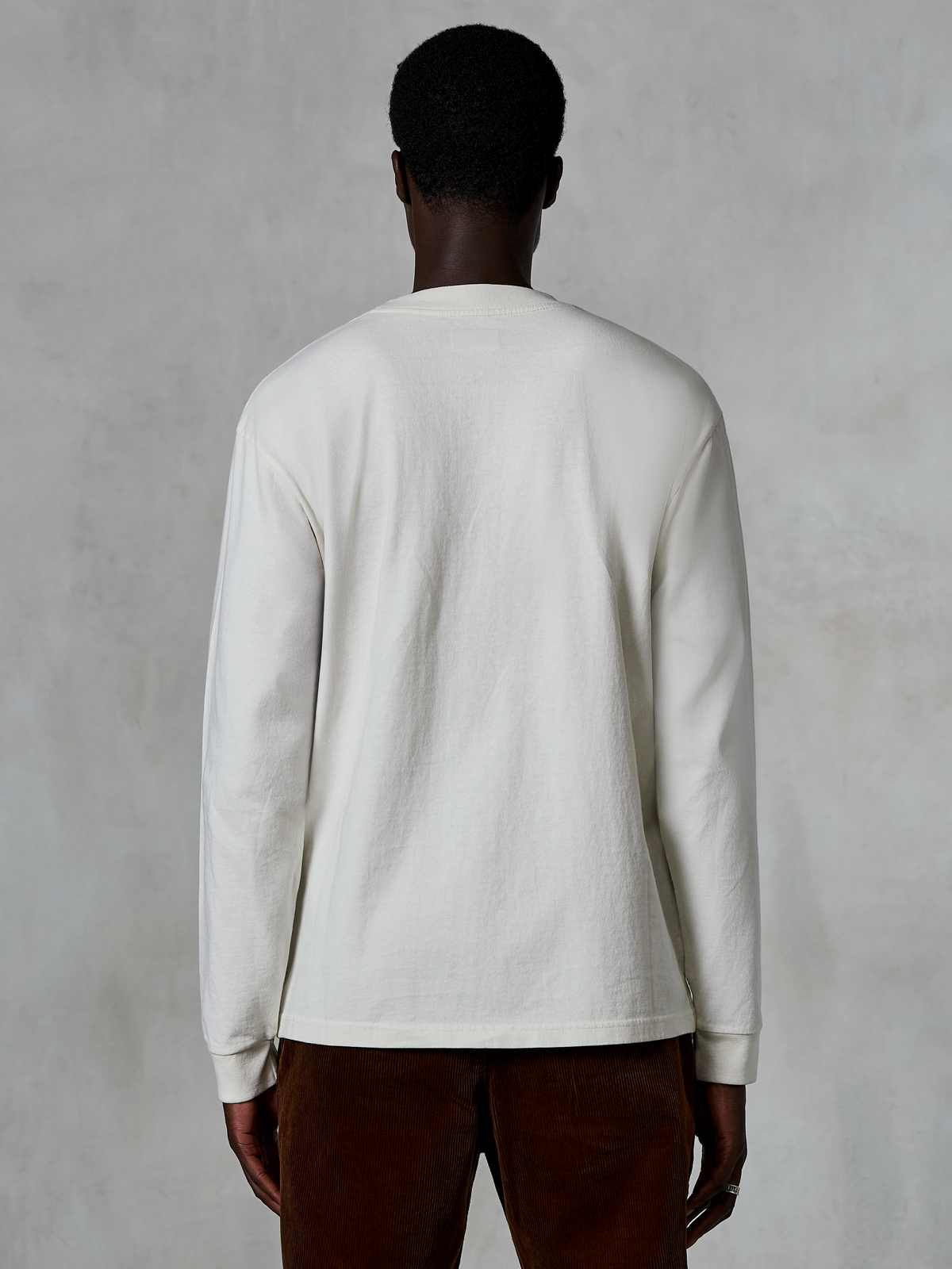 Alpinestars Essentials Ageless LS Tee, langærmet T-shirt, Off White, set bagfra på en person iført trøjen med brune fløjlsbukser, afslappet pasform, tekstureret økologisk bomuldsstof, minimalistisk afslappet streetwear