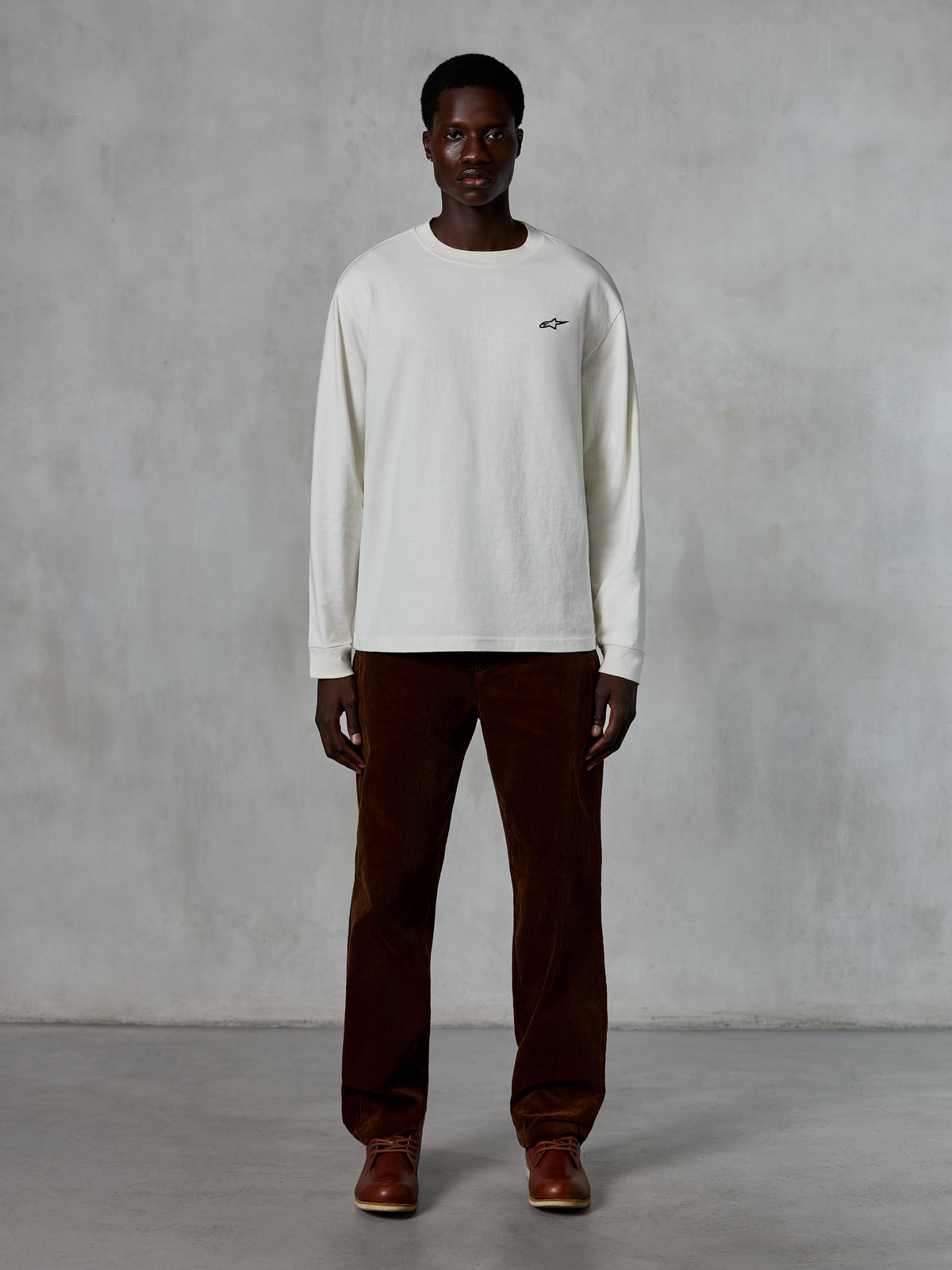 Alpinestars Essentials Ageless LS Tee, langærmet t-shirt, råhvid, båret af model med brune fløjlsbukser og brune læderstøvler, med et lille sort logo på brystet, afslappet moderne pasform, afslappet lifestyle streetwear