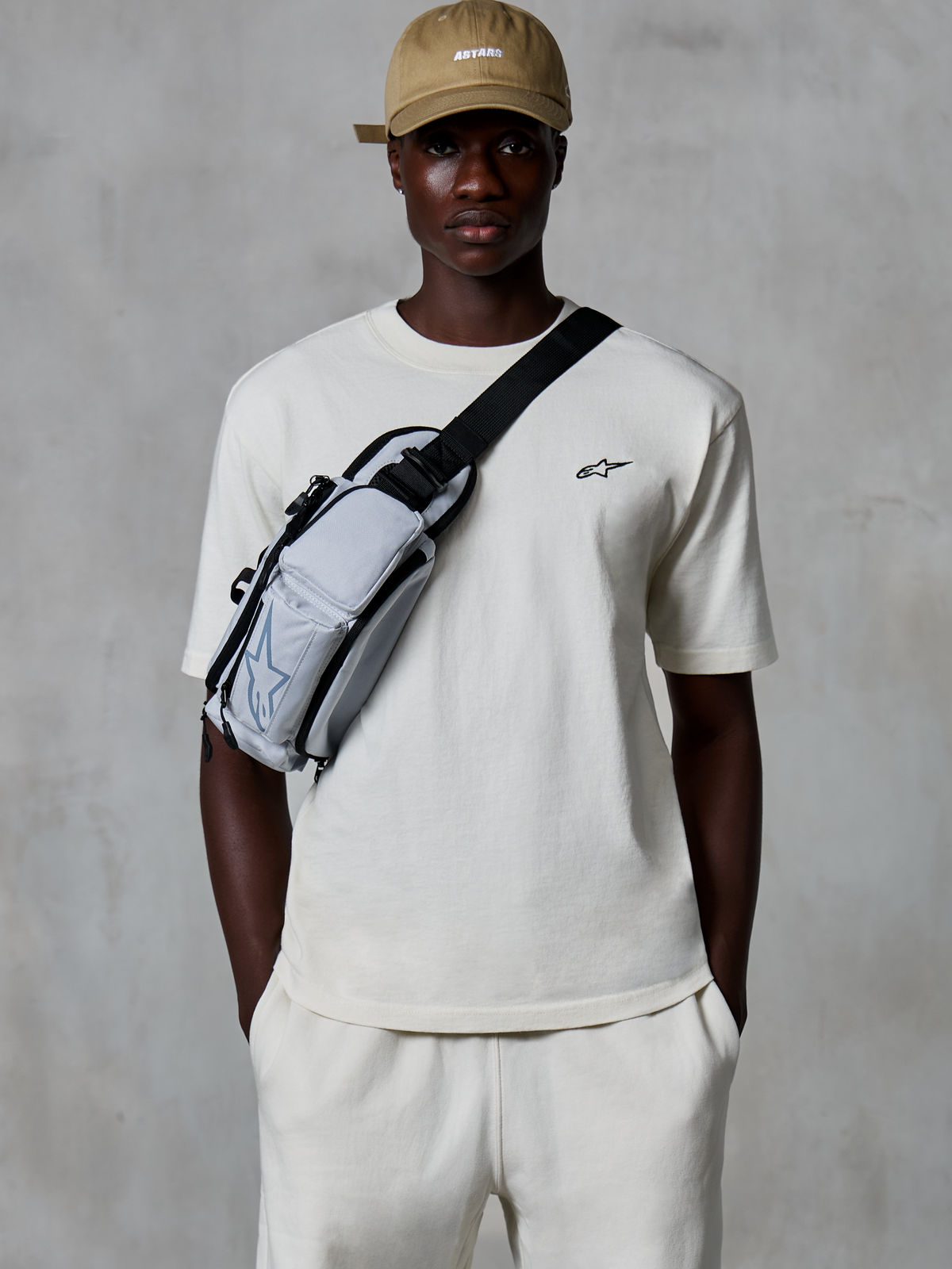 Alpinestars Essentials Ageless SS Tee, kortærmet T-shirt, råhvid, båret med matchende shorts og en sandfarvet kasket, med et lille sort logo på brystet og en grå crossbody-taske, afslappet motorsport-inspireret streetwear