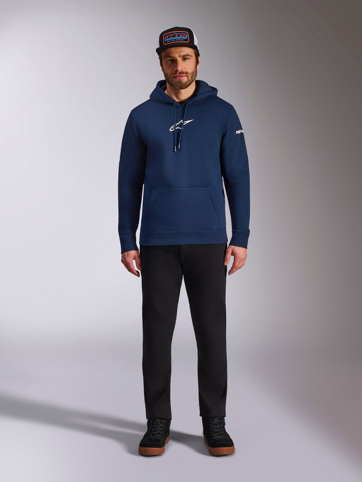 Male model wearing the Alpinestars Frontal Hoodie in Navy, pullover fleece with a white Astars logo on the chest and word logo on the sleeve, featuring a kangaroo pocket and drawstring hood, paired with black pants and a branded cap, casual motorsport streetwear