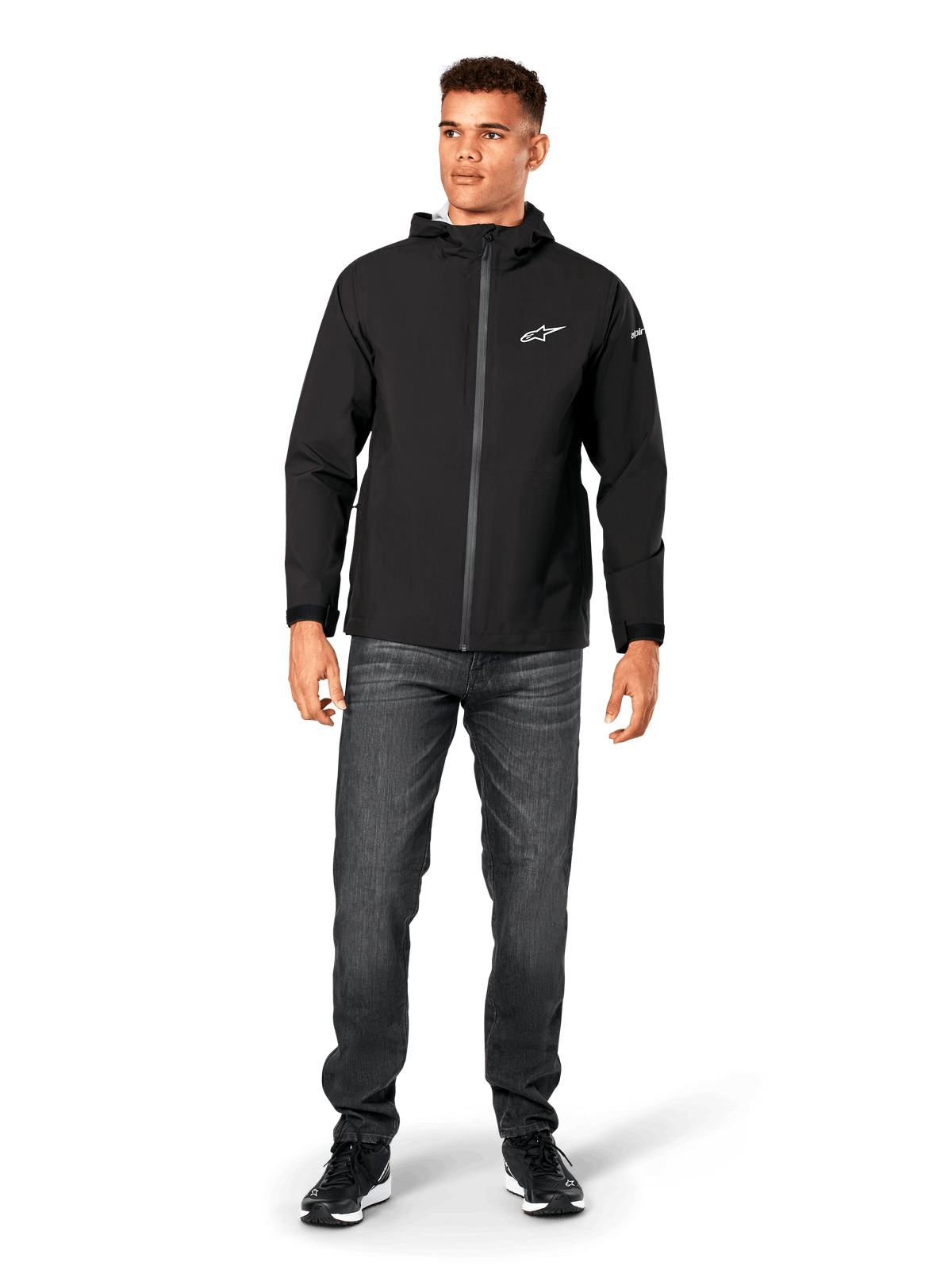 Alpinestars Kascade regnjakke, regnjakke til herrer, sort, båret med mørkegrå jeans og sorte sneakers, med hættedesign, belagt lynlås foran og hvidt Alpinestars-logo på brystet, afslappet motorsport-inspireret streetwear