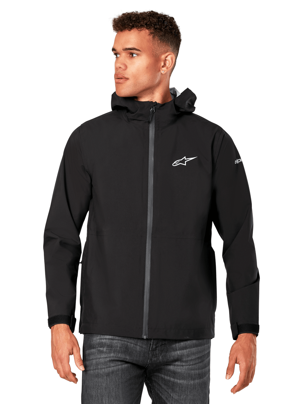 Alpinestars Kascade regnjakke, mandlig model i sort regnjakke med hætte, hvidt brystlogo og branding på ærmet, belagt lynlås foran og velcro-manchetter, mørke jeans, afslappet motorsport-inspireret streetwear