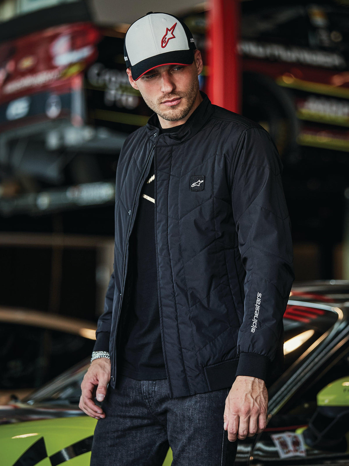 Alpinestars Precedent jakke, sort quiltet jakke med Formel 1-styling, med et ikonisk vinklet quiltmønster, logomærke på brystet og hvidt broderi på ærmet, båret med en matchende Alpinestars cap i et motorsport-garage-miljø.
