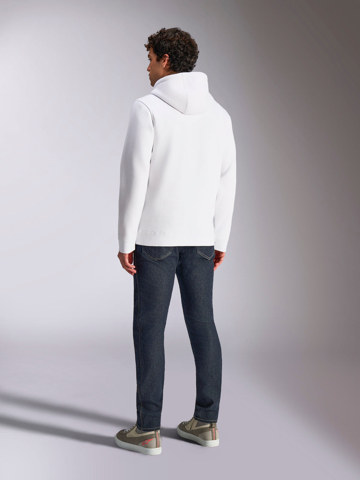 Alpinestars Blaze V3 Hættetrøje, mandlig model set bagfra iført hvid pullover-hættetrøje, afslappet sportstøj stylet med mørkeblå jeans og grå sneakers, moderne motorsport-inspireret design