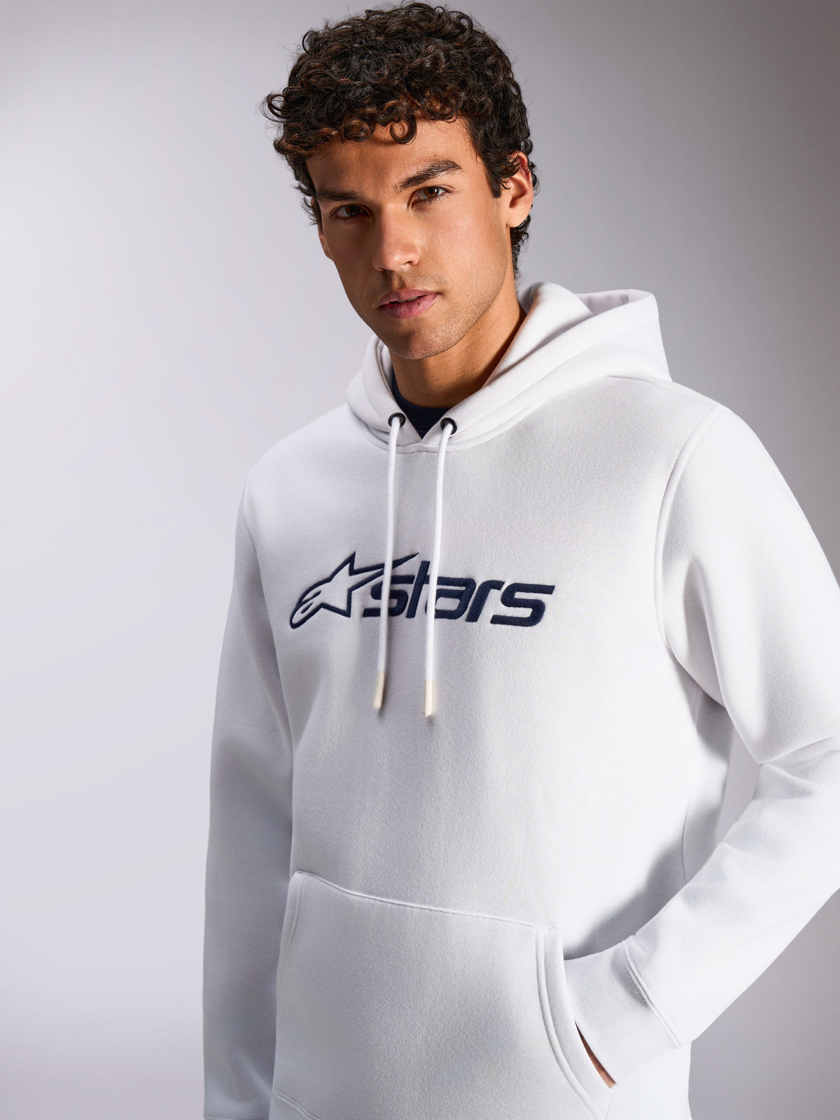 Alpinestars Blaze V3 hættetrøje, hvid og navy, båret af mandlig model, afslappet sportstøj, mørkeblåt Alpinestars-logo på fronten, hætte med snøre, kængurulomme, moderne livsstilsdesign