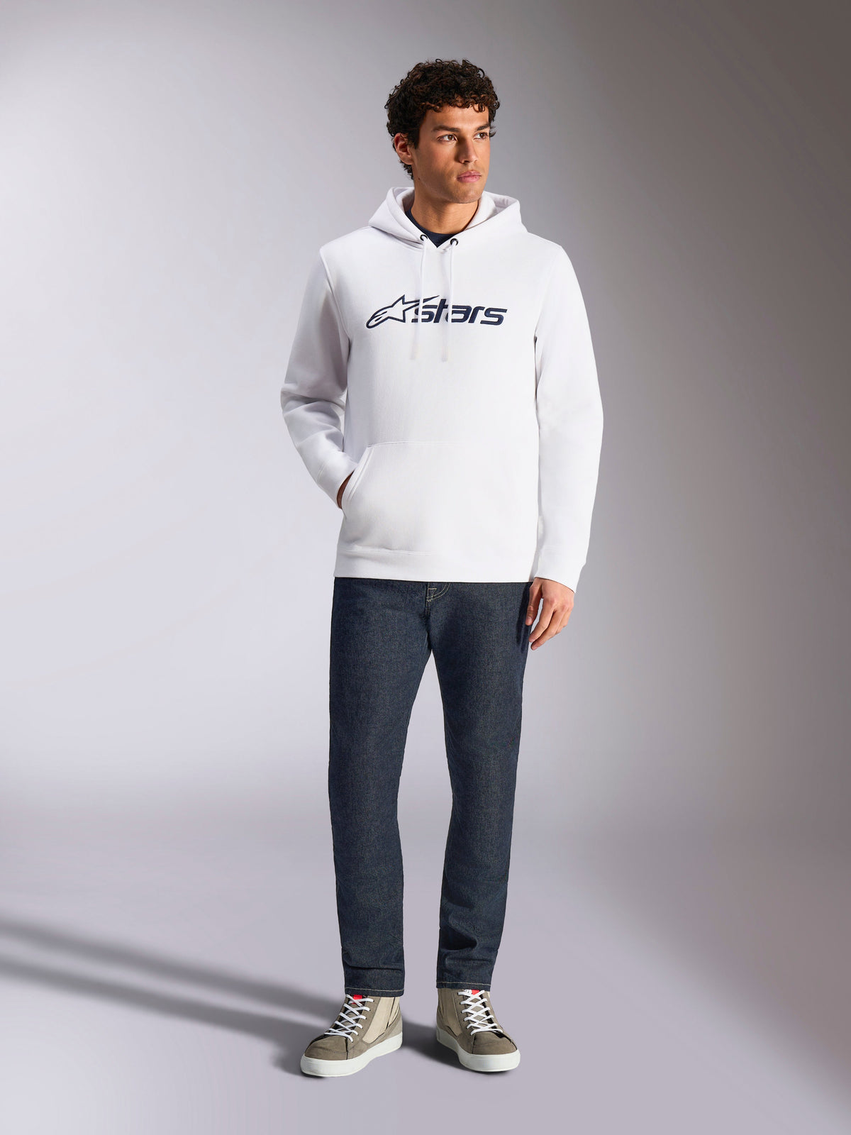 Alpinestars Blaze V3 hættetrøje, hvid afslappet streetwear-hættetrøje med mørkeblåt Alpinestars-logo på brystet, båret af en model med mørkeblå jeans og sandfarvede sneakers, med hætte med snoretræk og kængurulomme på fronten