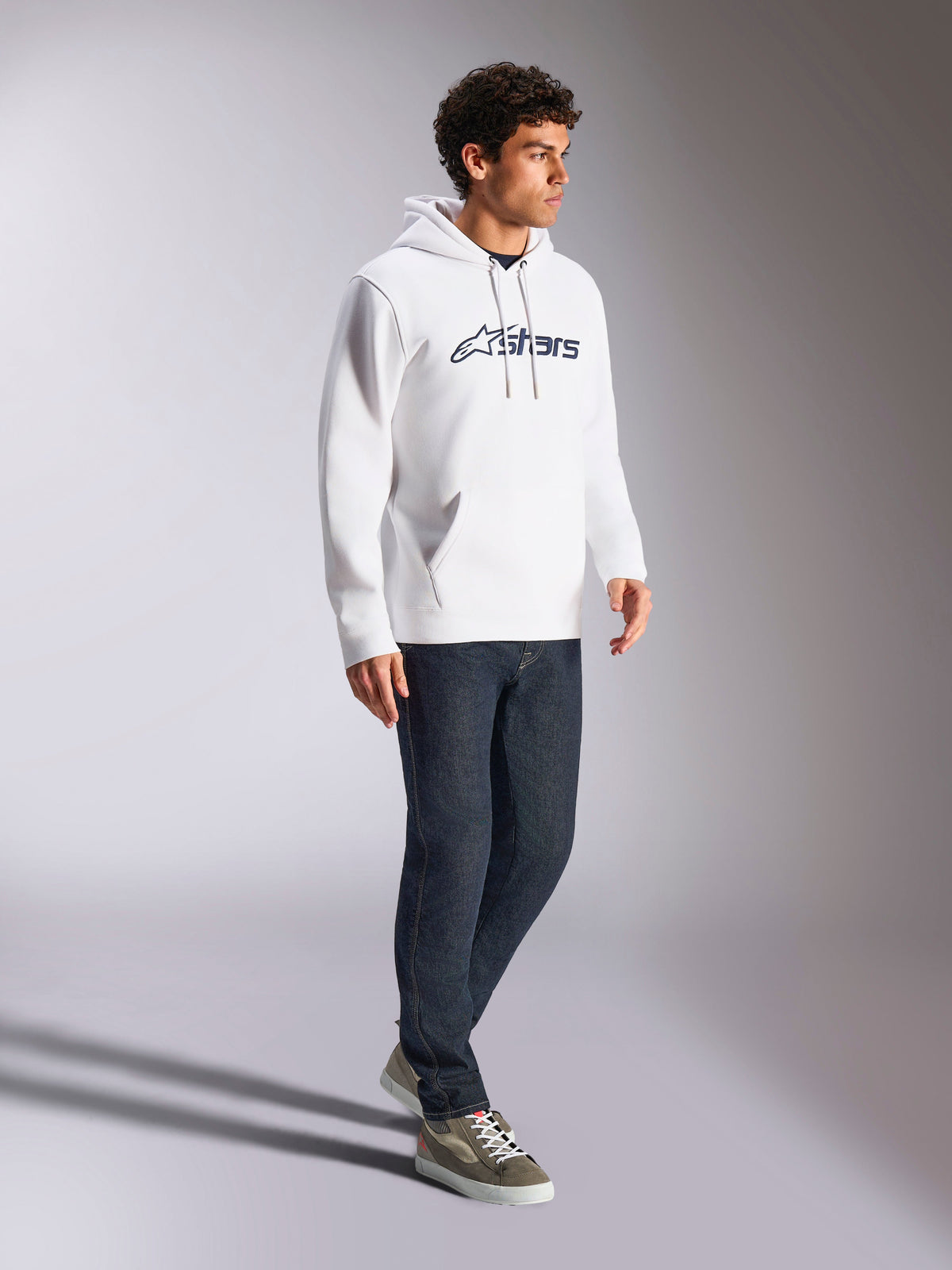 Alpinestars Blaze V3 hættetrøje, hvid og navy, afslappet streetwear båret af en model med mørkeblå jeans og sneakers, har hætte med snoretræk og markant logoprint på fronten, moderne motorsport-inspireret design