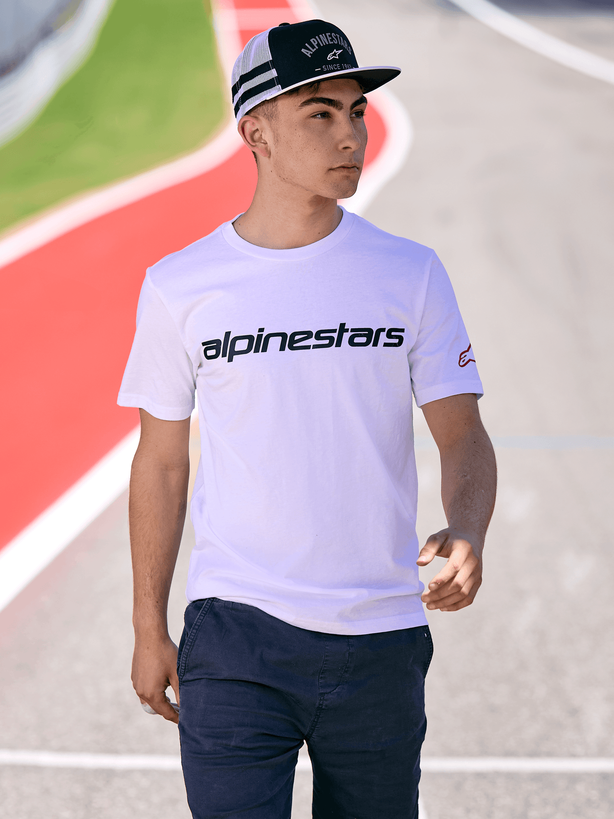 Mand iført Alpinestars Backline Trucker Hat i sort og hvid, kombineret med en hvid Alpinestars logo t-shirt og mørkeblå bukser, casual motorsport streetwear på en udendørs racerbane-baggrund